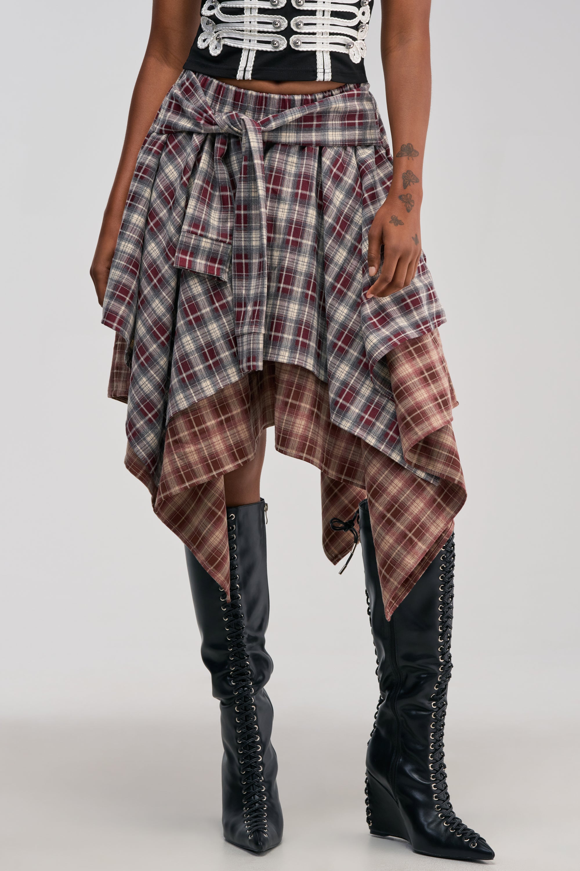 Wearing the FEELING ALL OVER THE PLACE PLAID MIDI SKIRT—a layered, handkerchief hem piece in striking red and gray plaid—paired with a black and white top and black lace-up knee-high boots, this look delivers edgy streetwear vibes perfect for a concert outfit. A tattoo on the wearer’s hand adds to the cool, rebellious aesthetic.