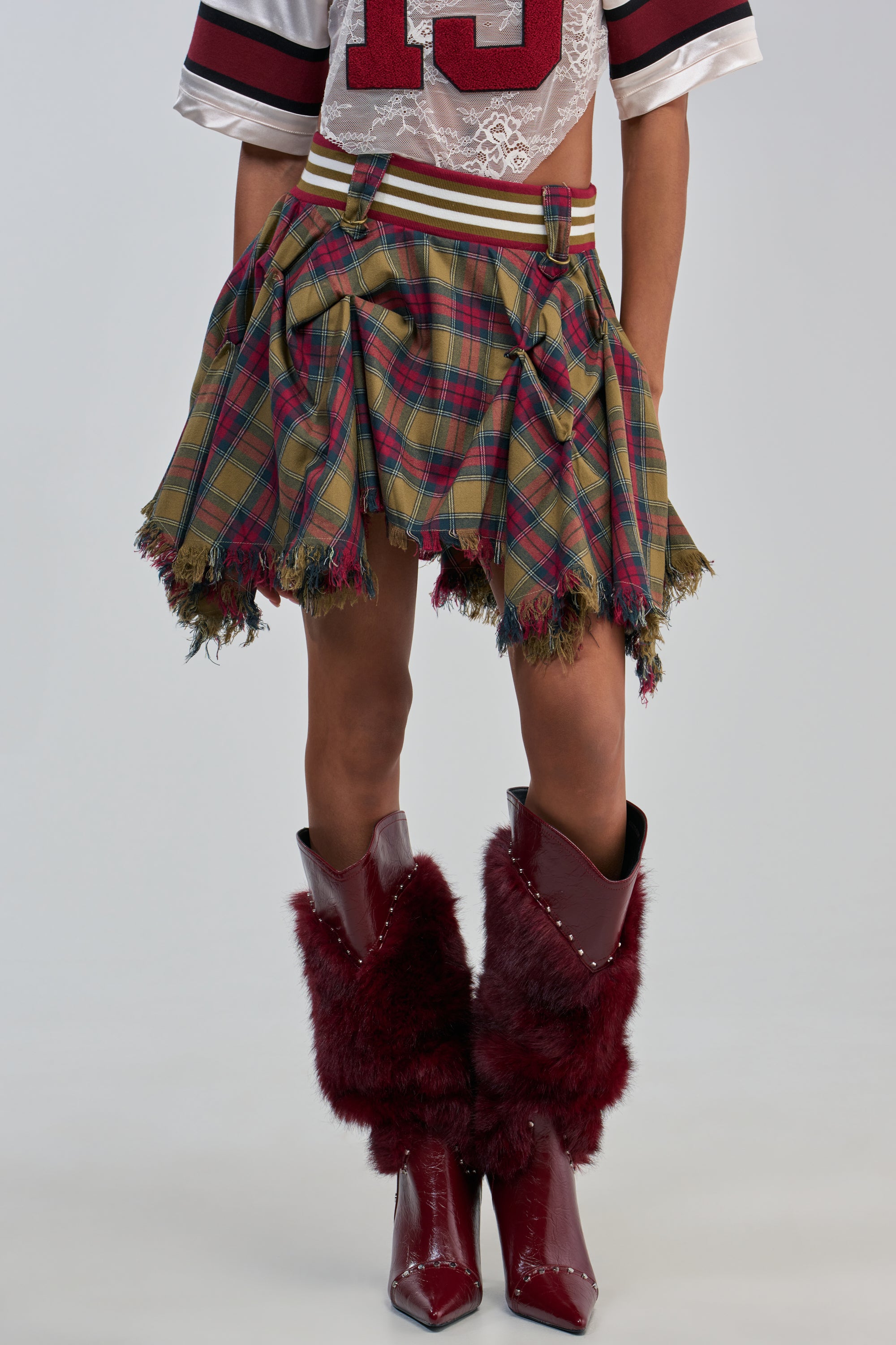 Wearing the CRAZY FOR YOU PLAID MINI SKIRT with frayed edges, a sporty jersey crop top, and tall burgundy furry cowboy boots, this look effortlessly captures cool preppy streetwear vibes. The background is plain and light-colored, with only the lower body visible.