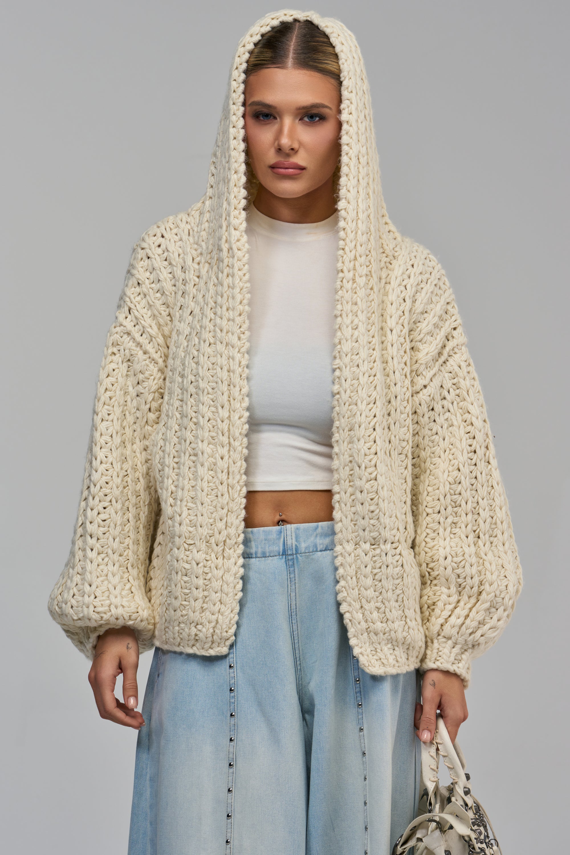 A person wearing a COZY CROCHET HOODED CARDIGAN IN IVORY over a white crop top and light-wash jeans.