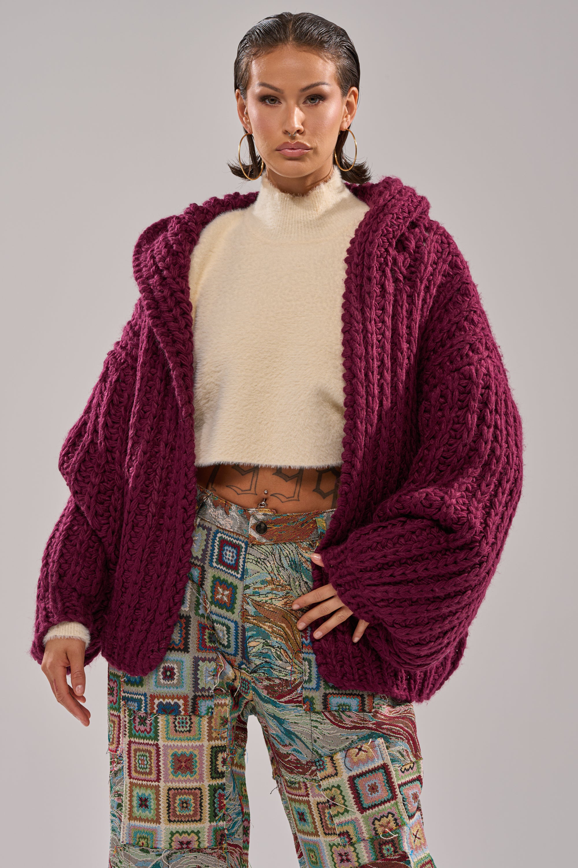 A woman stands confidently against a neutral background wearing the COZY CROCHET HOODED CARDIGAN in maroon over a cream turtleneck sweater and patterned pants with geometric and animal prints.