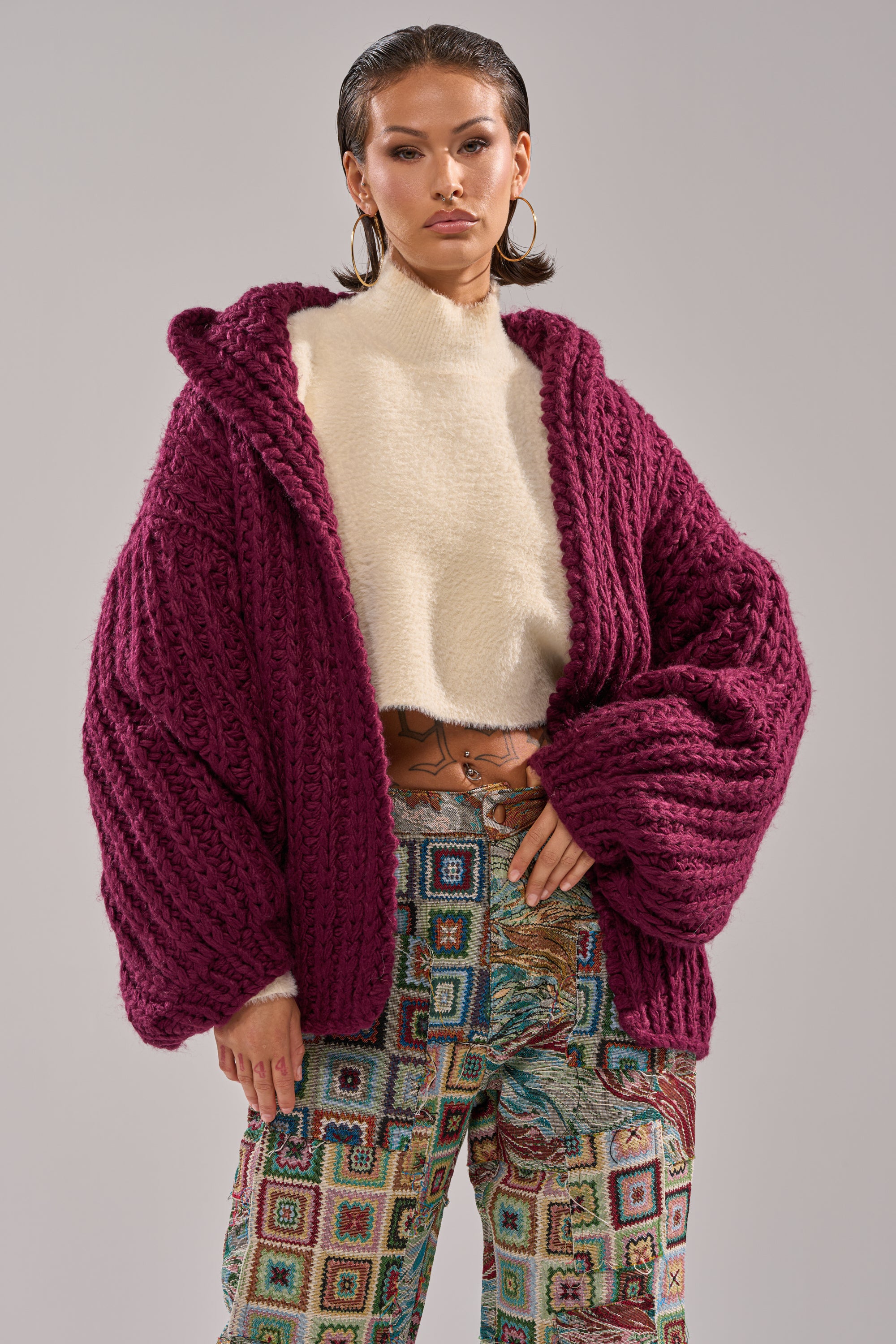 A person wears the COZY CROCHET HOODED CARDIGAN over a cream fuzzy sweater and colorful patterned pants, standing confidently against a plain background.