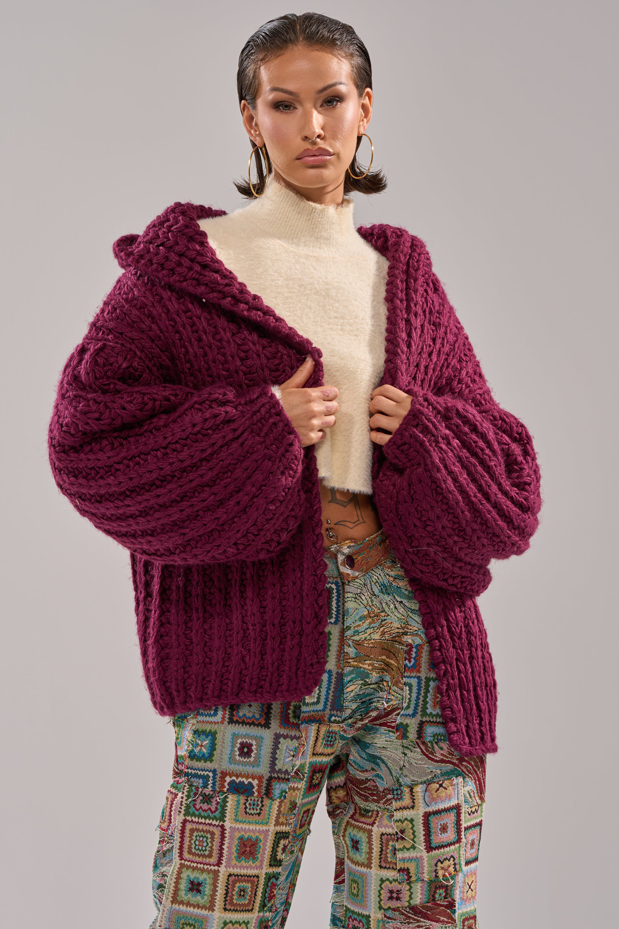 A woman models the COZY CROCHET HOODED CARDIGAN in deep magenta over a cream turtleneck, paired with vibrant geometric and floral patterned trousers, standing against a plain gray backdrop.