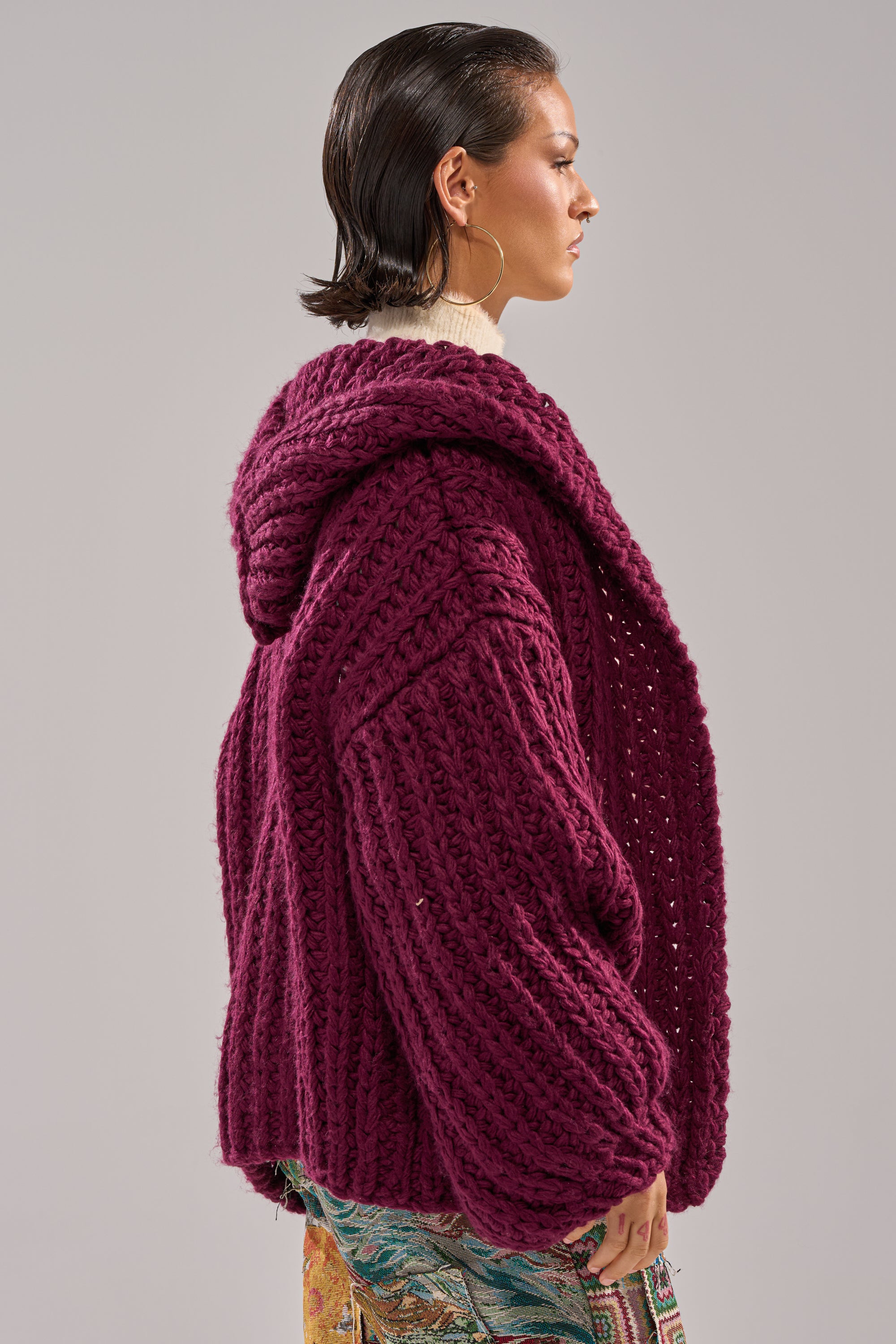 A person with short, slicked-back hair models the COZY CROCHET HOODED CARDIGAN in deep purple, featuring a chunky hand-knit design and large hood. They stand sideways against a plain light grey background.