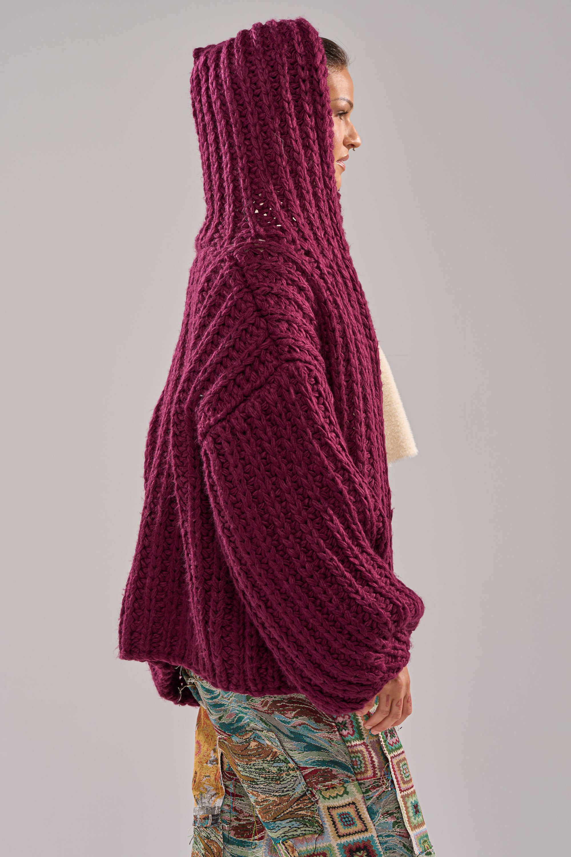 A person in profile wears the COZY CROCHET HOODED CARDIGAN in deep magenta, styled with multicolored patterned pants against a plain, light background.