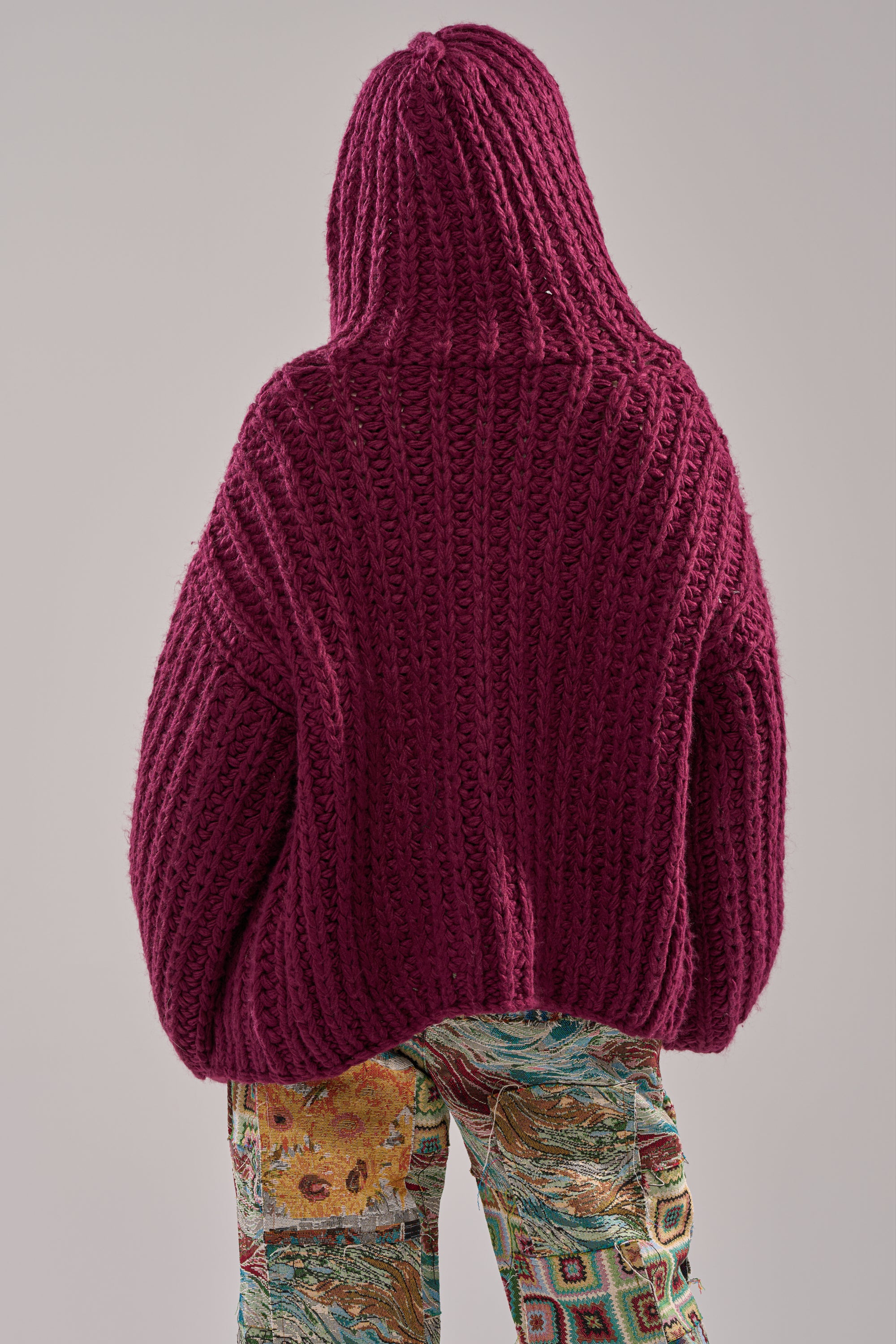 An individual faces away from the camera, wearing the COZY CROCHET HOODED CARDIGAN in vibrant magenta with colorful, patterned pants, set against a neutral background.