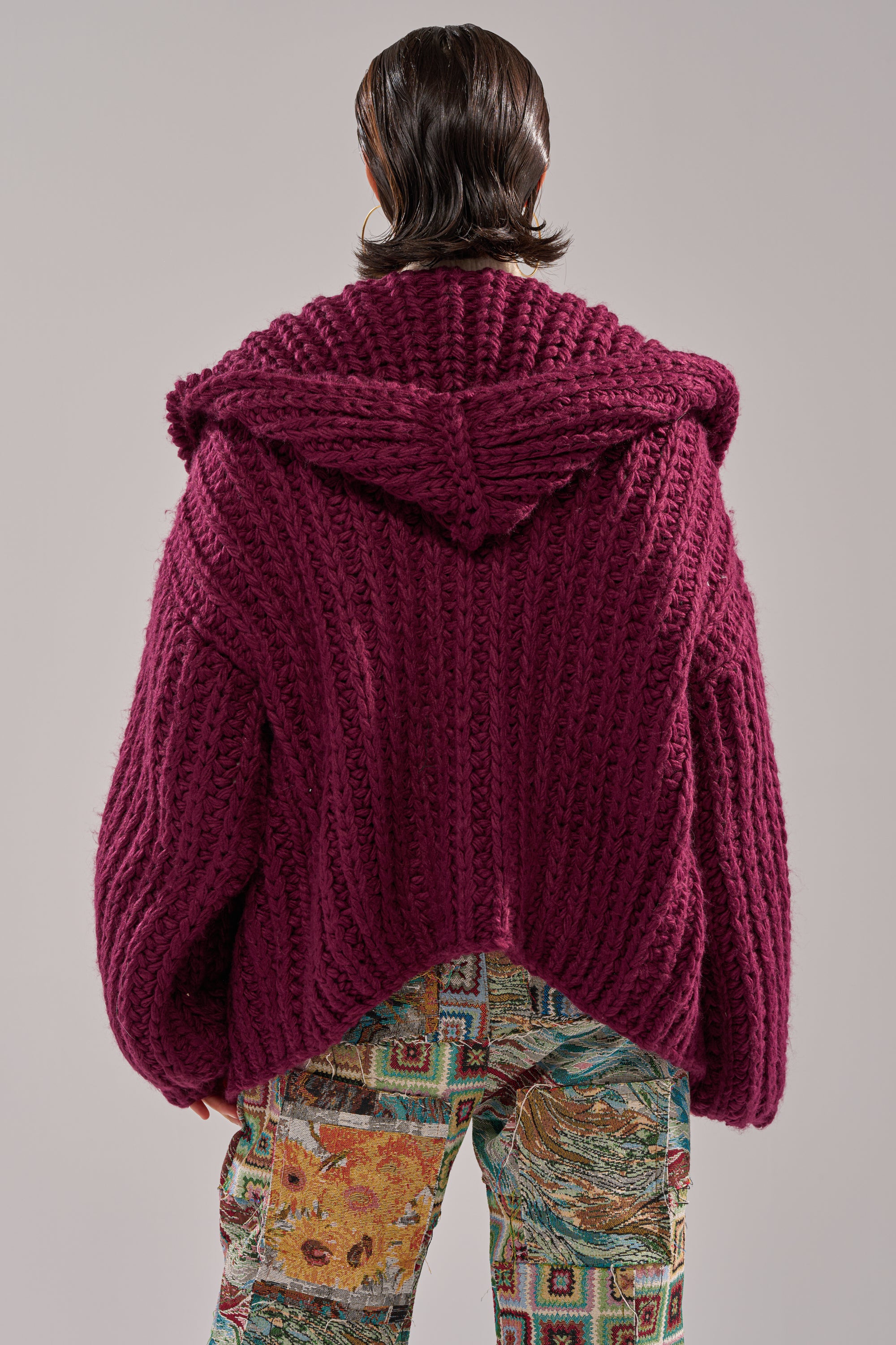 A person with straight, dark, shoulder-length hair is seen from behind, wearing the COZY CROCHET HOODED CARDIGAN in deep magenta and colorful patterned pants featuring various designs and textures.
