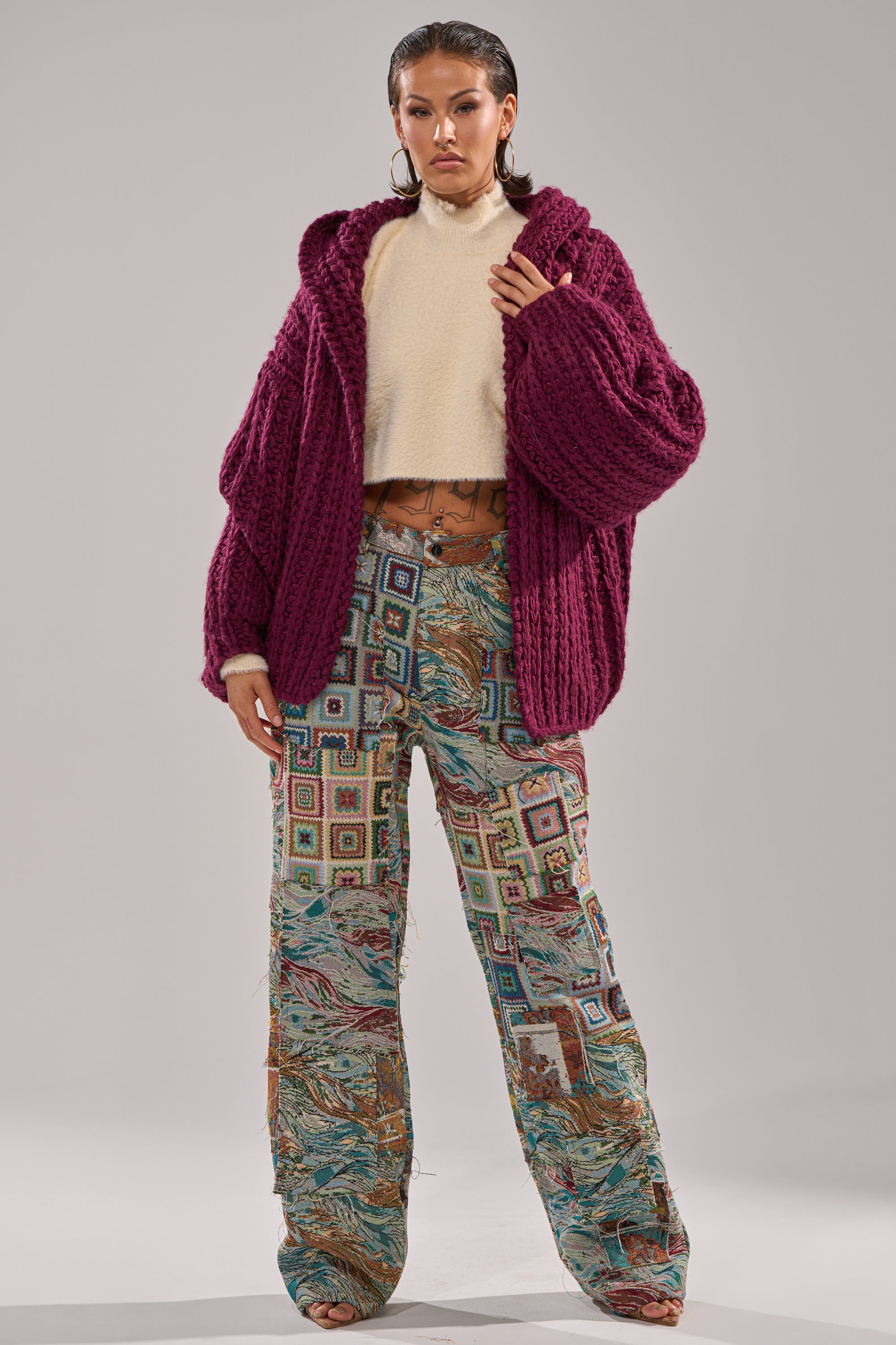 A woman stands confidently in the COZY CROCHET HOODED CARDIGAN, layered over a cream turtleneck and loose, patterned patchwork pants. She pairs the look with wet-look, slicked-back hair and hoop earrings.