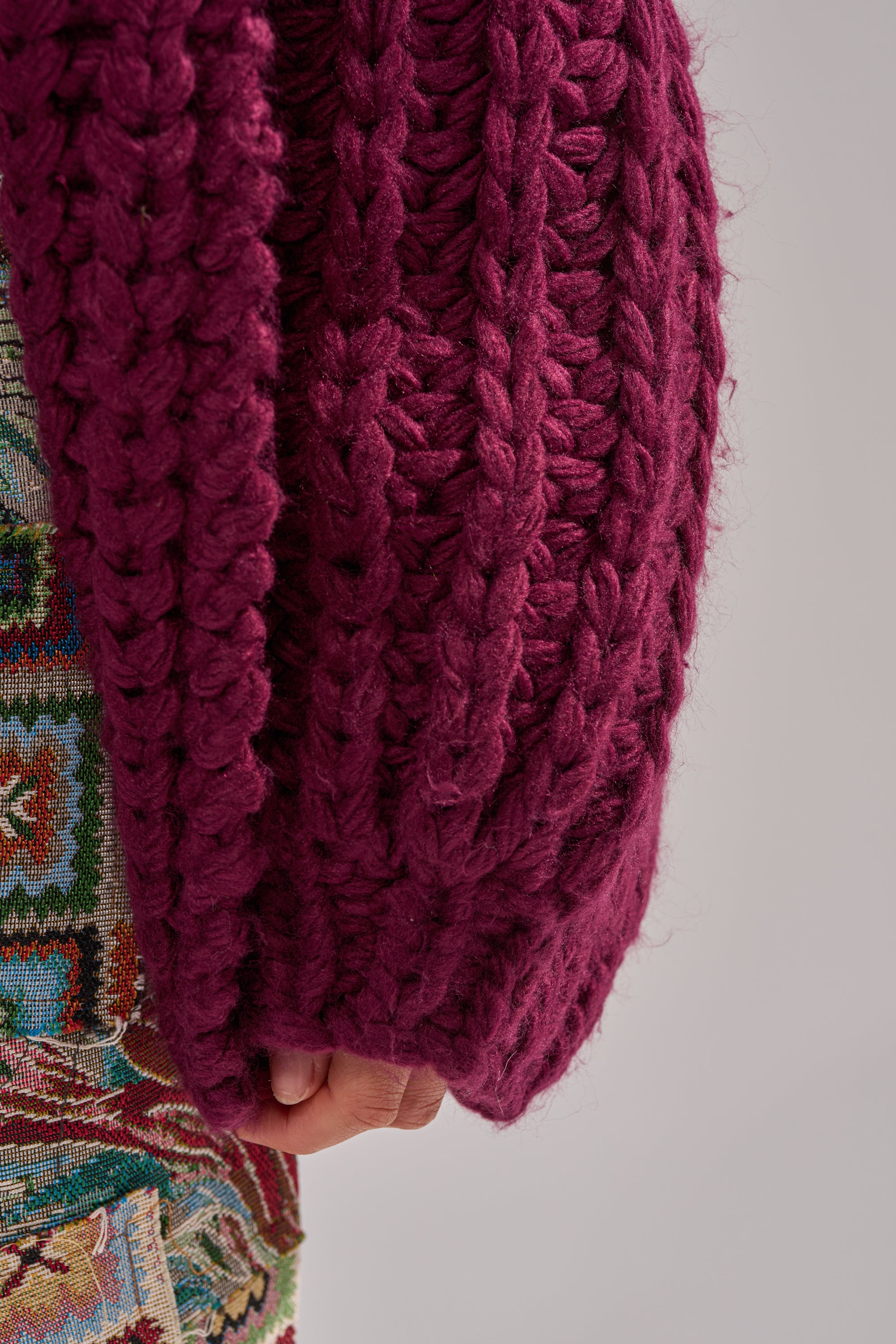 A close-up shows a hand partly covered by the sleeve of a COZY CROCHET HOODED CARDIGAN in deep magenta, beside vibrant patterned fabric on a neutral background.