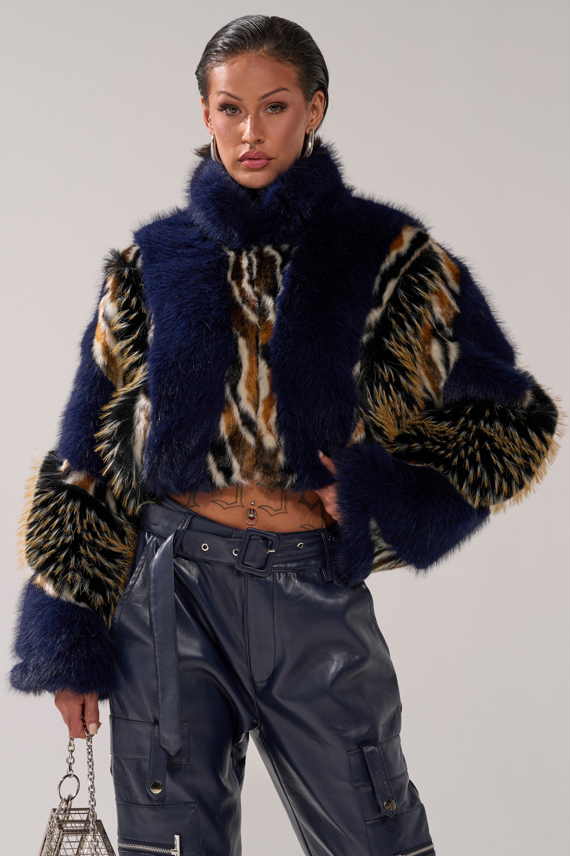 A woman poses confidently against a plain background in the CYCLONE PATCHWORK FAUX FUR—a bold, cropped jacket with tiered fur striping and animal print patterns—paired with shiny leather pants and a small silver handbag.