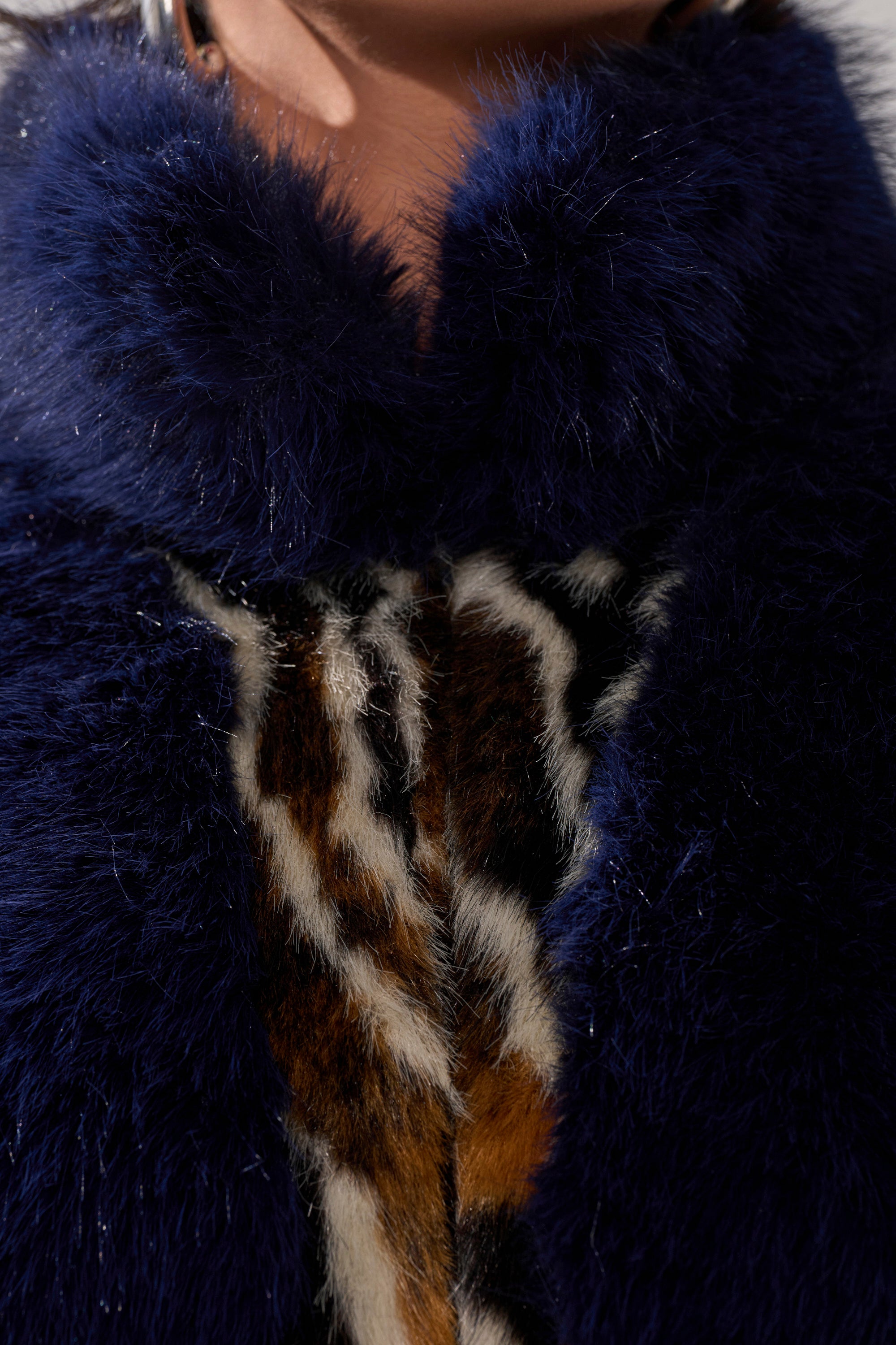 Close-up of a person wearing the CYCLONE PATCHWORK FAUX FUR jacket in dark blue with a collar, revealing a section of brown, black, and white patterned patchwork faux fur underneath.