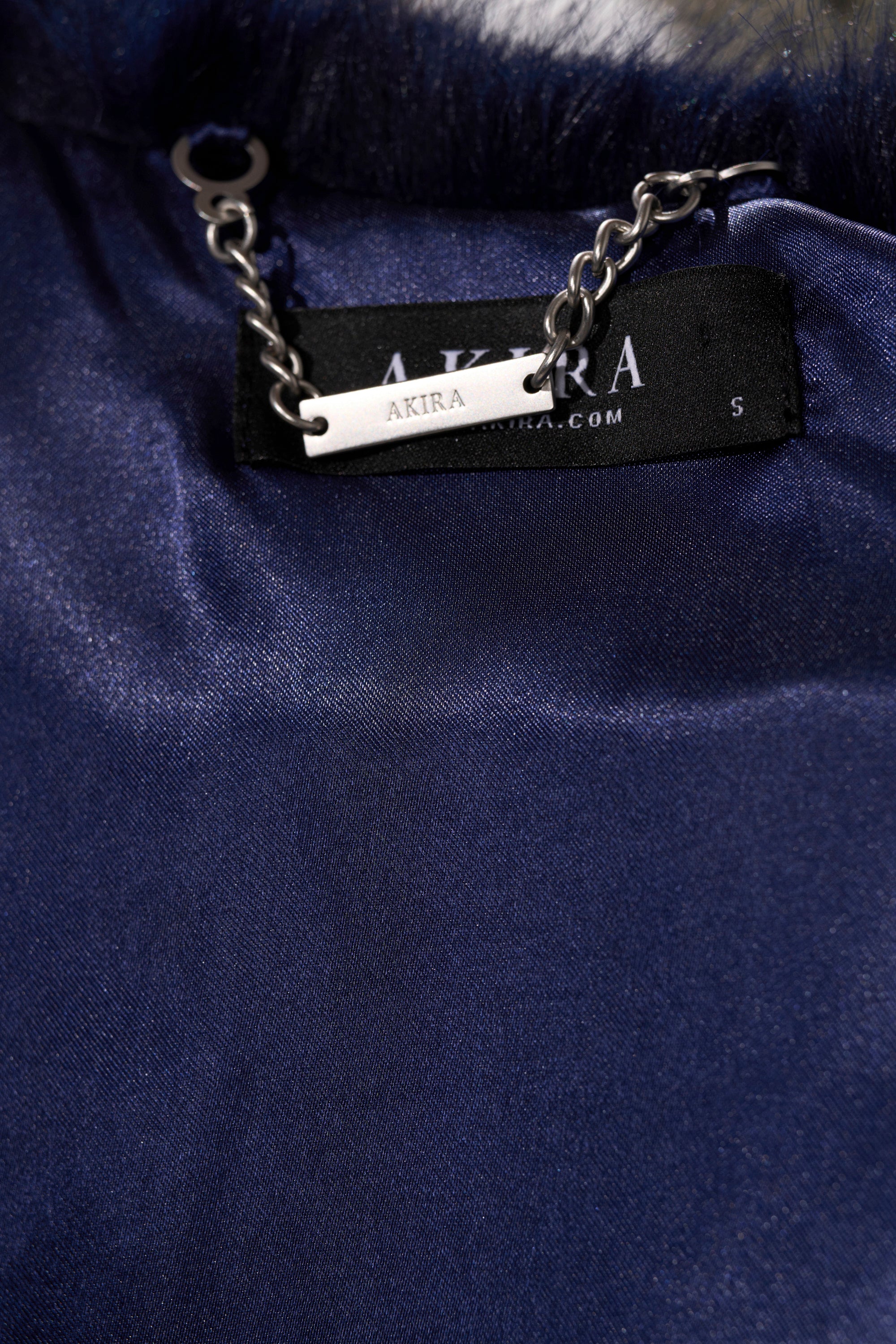 Close-up of dark blue satin lining inside the CYCLONE PATCHWORK FAUX FUR jacket, with a black "AKIRA" label, metal chain above it, and a tag showing size "S".