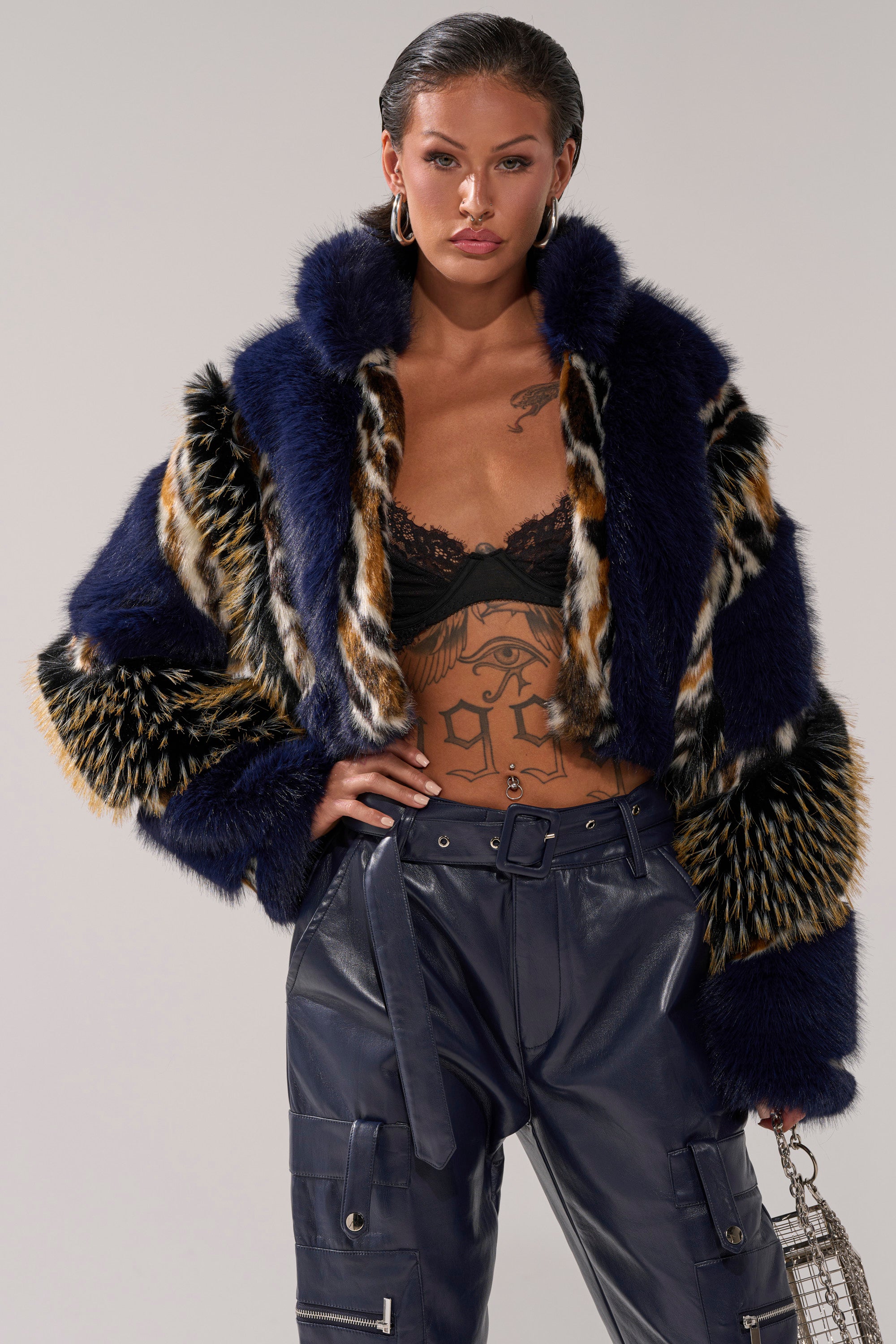 A woman wears the CYCLONE PATCHWORK FAUX FUR jacket featuring dark blue and animal prints, paired with a black lace bralette, navy high-waisted cargo pants, hoop earrings, and a small metallic handbag.