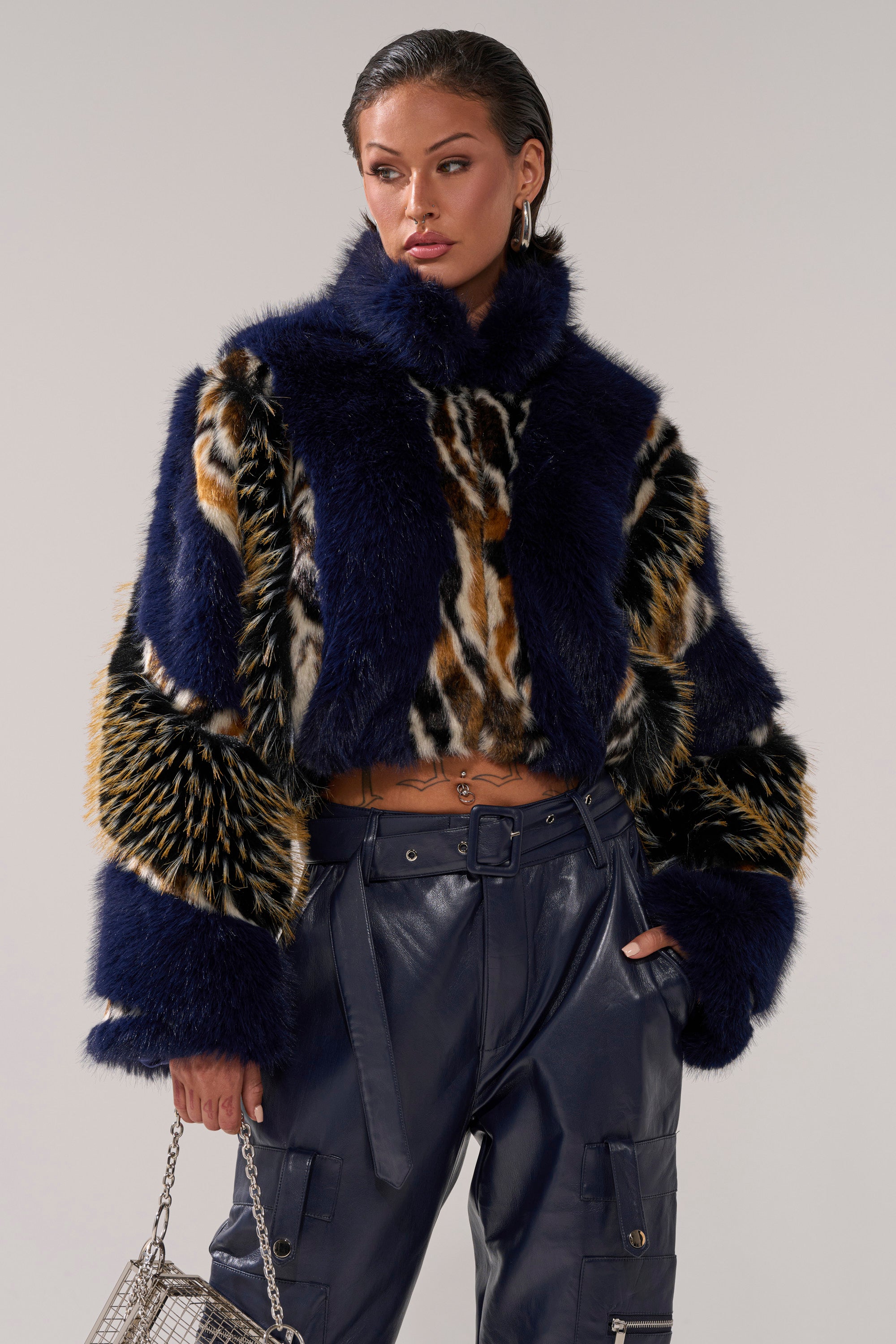 A woman models the CYCLONE PATCHWORK FAUX FUR jacket with animal prints, shiny navy high-waisted pants, and a small silver handbag, posing confidently with one hand in her pocket.