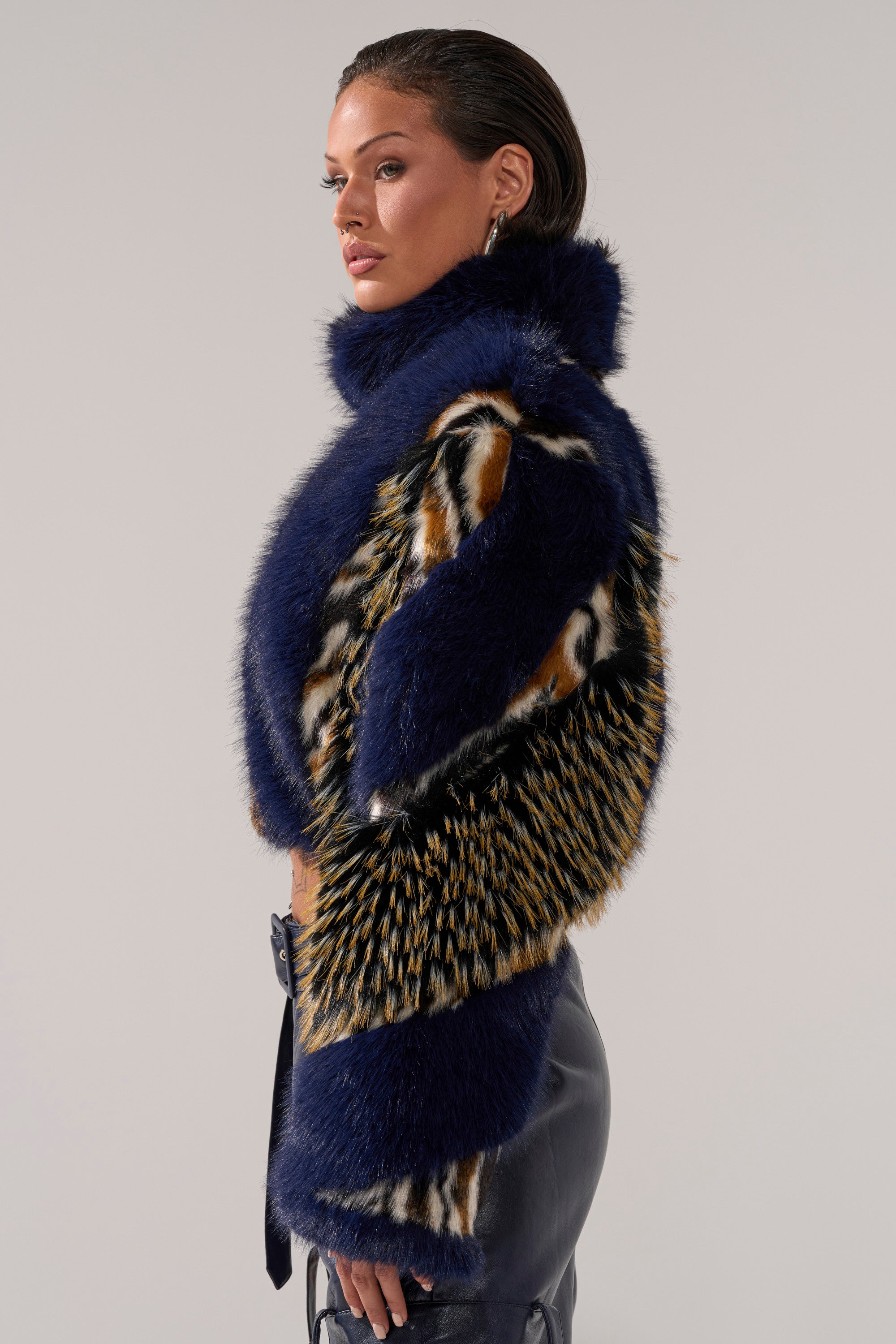A woman stands in profile wearing the CYCLONE PATCHWORK FAUX FUR jacket in blue, black, and tan tones with a high collar, paired with black leather pants against a plain light background.