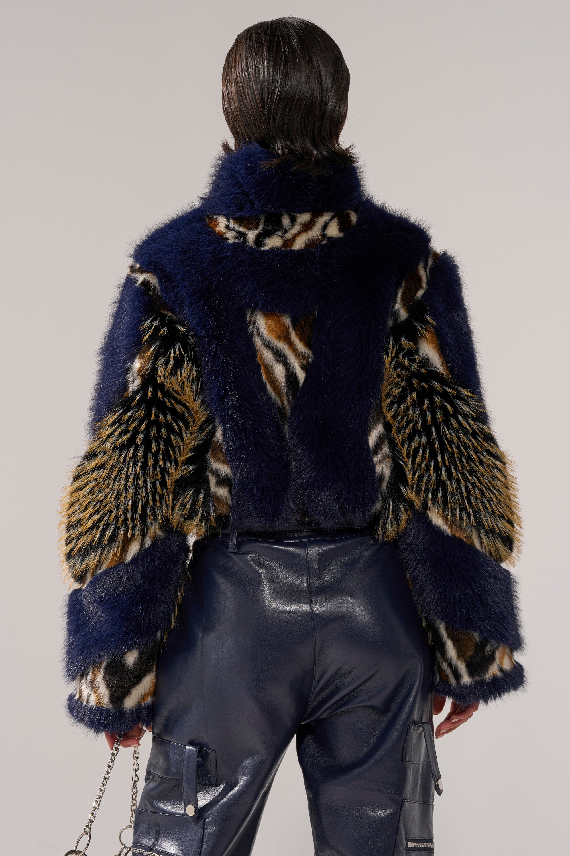 A person with slicked-back hair is seen from behind wearing the CYCLONE PATCHWORK FAUX FUR jacket—blue, with animal print and textured sleeves—paired with shiny dark blue pants against a plain background.