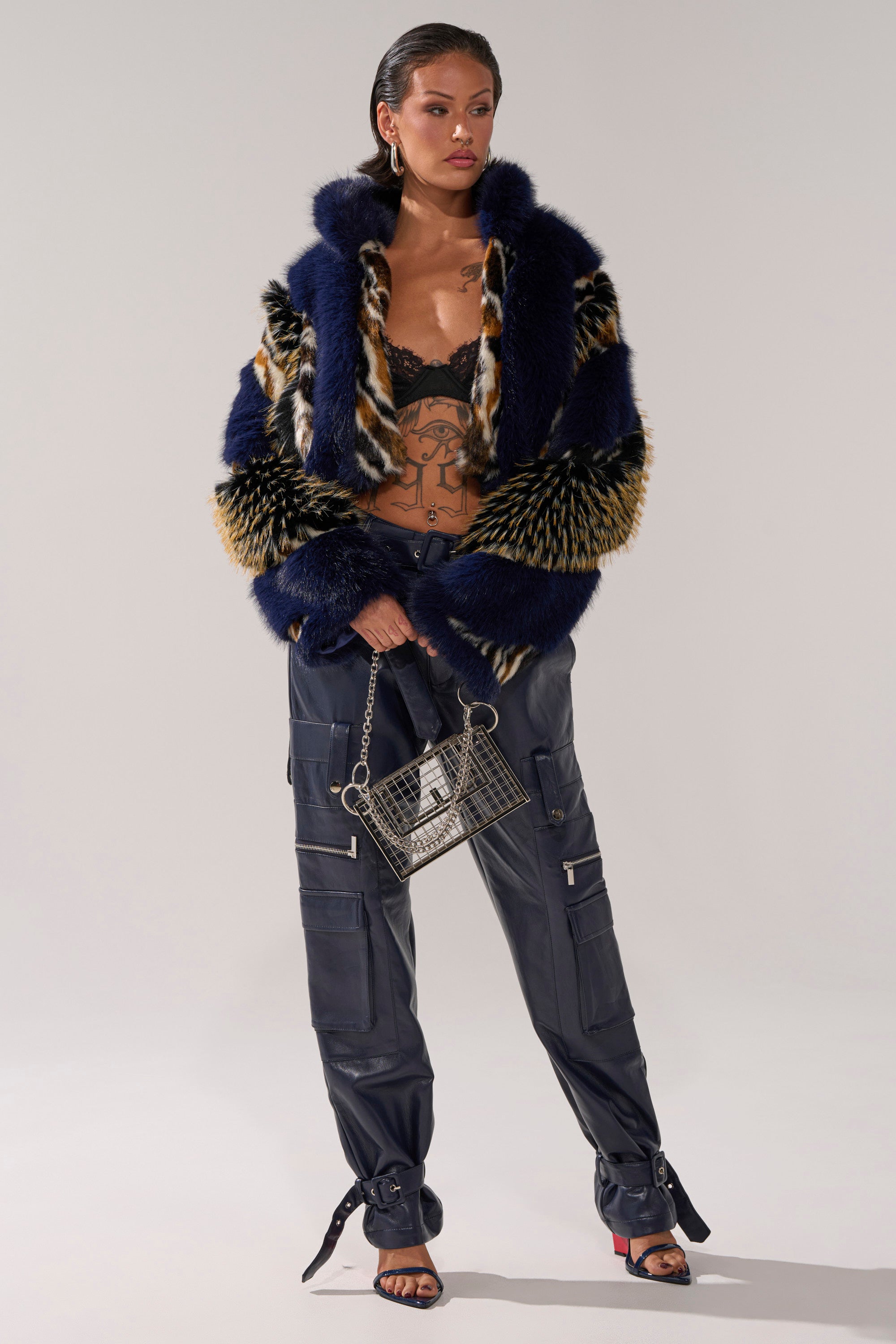 A woman poses in the CYCLONE PATCHWORK FAUX FUR jacket over a black lace bralette, navy cargo pants, black heeled sandals, hoop earrings, and carries a metallic mesh handbag. Tattoos adorn her torso.