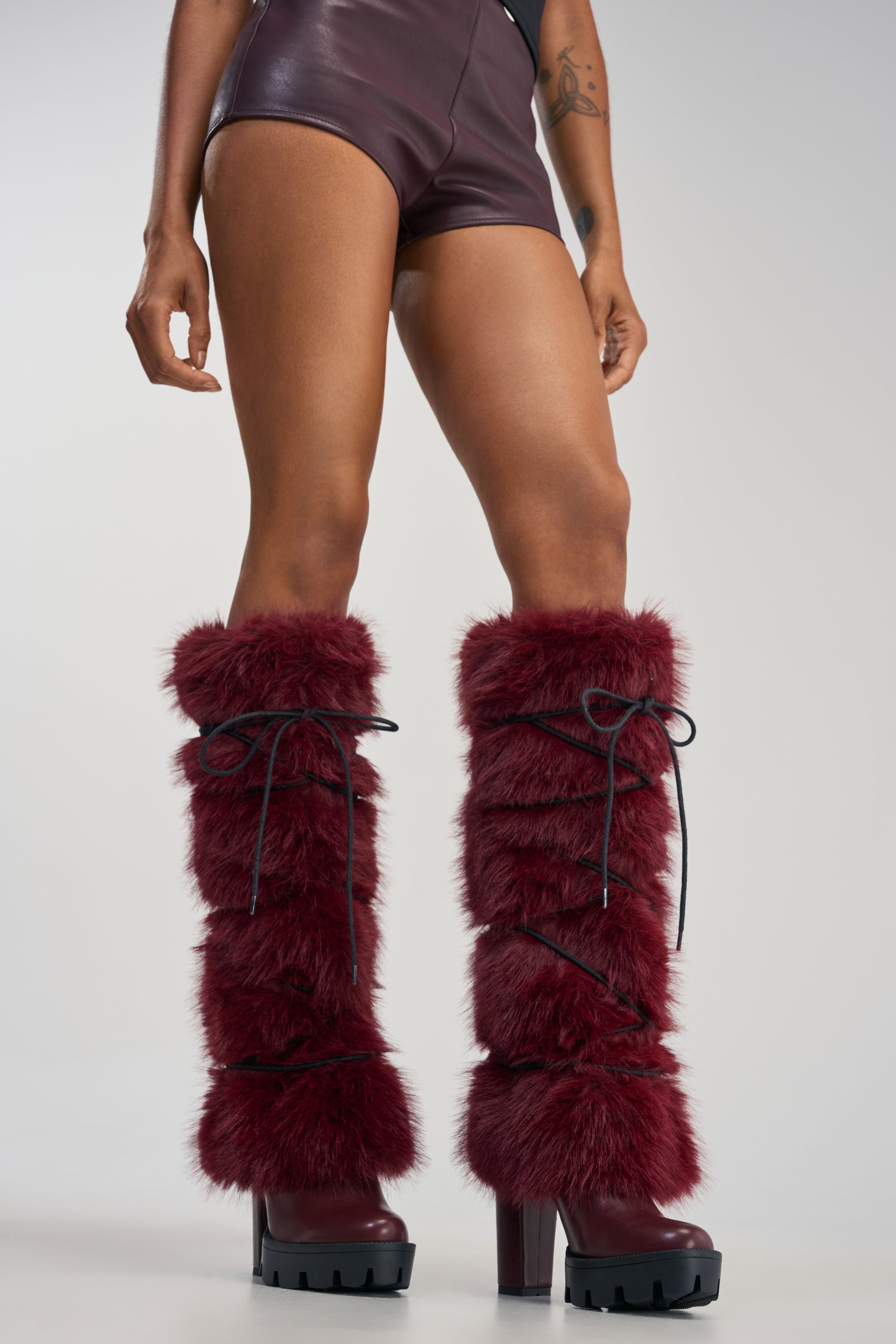A person wearing dark shorts and the AZALEA WANG CYRENE BURGUNDY FUR BLOCK HEEL BOOT with striking knee-high burgundy faux fur and black laces stands against a plain background.