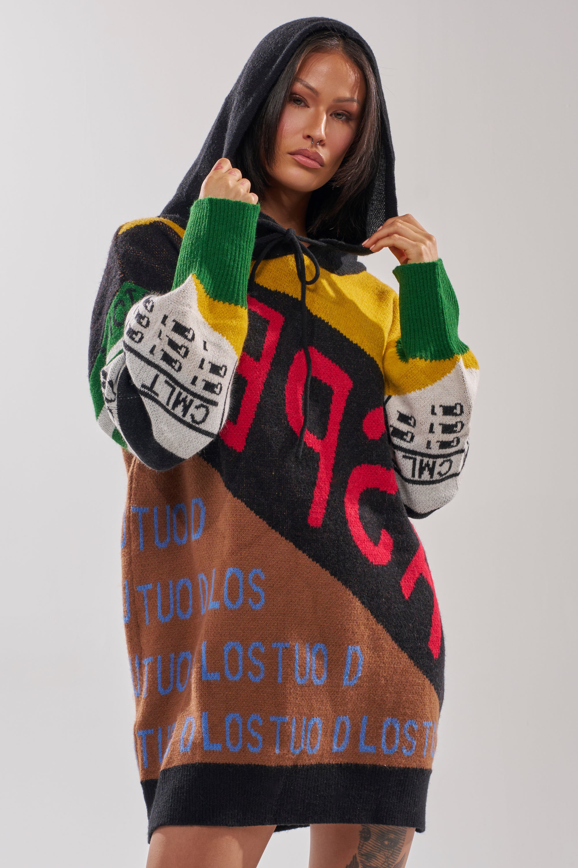 A woman faces forward in the ALPHA FEMALE HOODED SWEATSHIRT DRESS IN YELLOW MULTI, featuring bold patterns, large letters, and graphic text in vibrant red, blue, green, yellow, and black. Hood up, she holds the drawstrings for a true streetwear vibe.