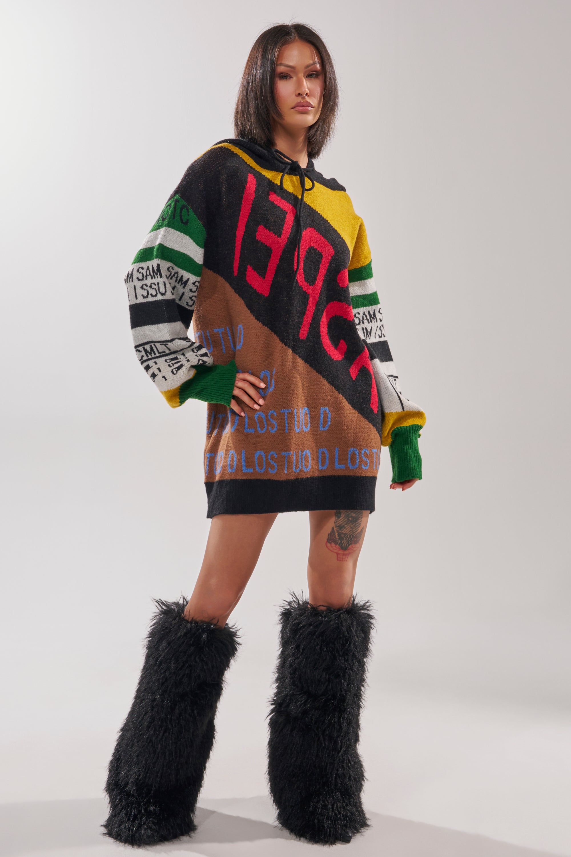 A woman with a bob haircut confidently poses in the ALPHA FEMALE HOODED SWEATSHIRT DRESS IN YELLOW MULTI, paired with black furry knee-high boots, against a plain light background.