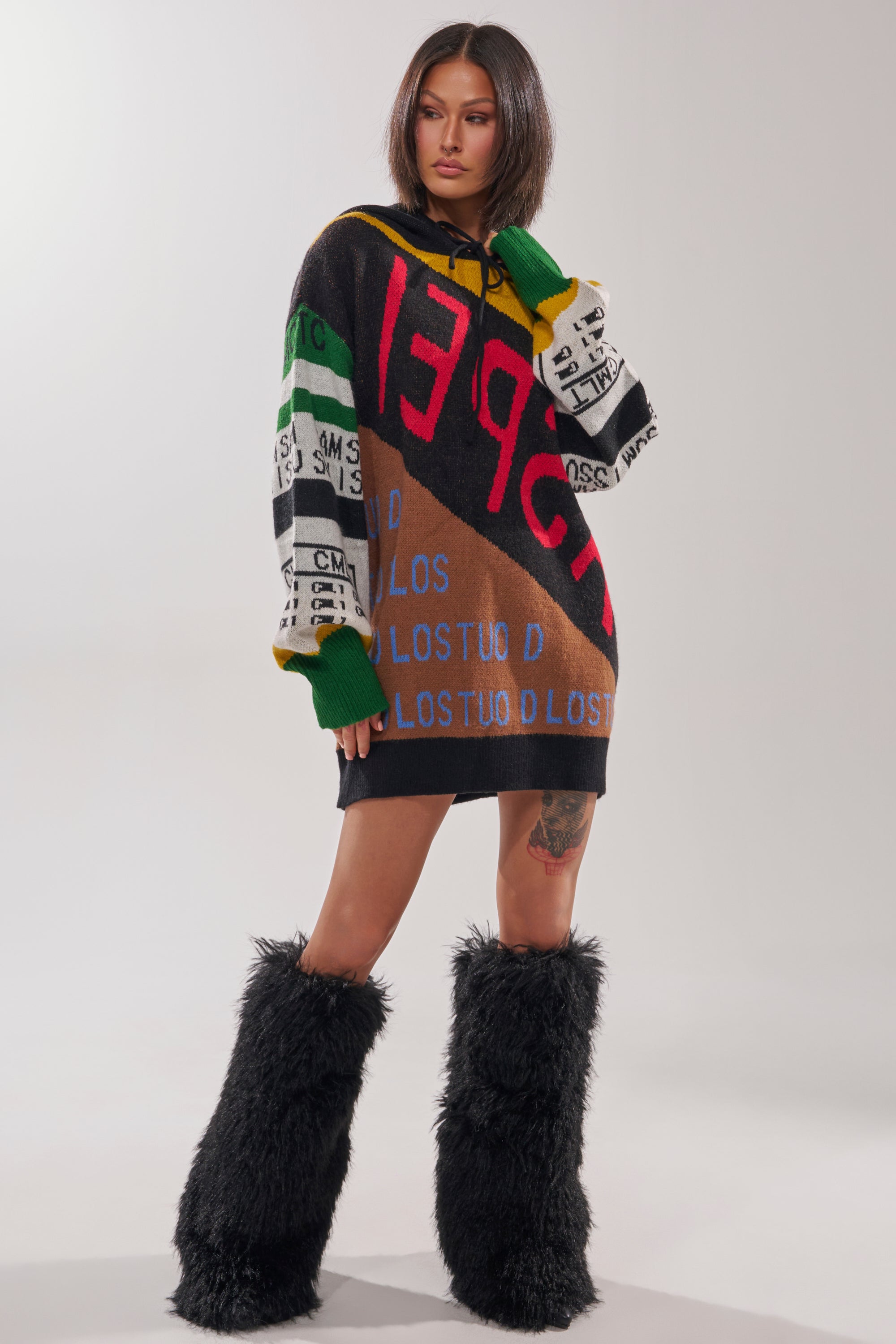 A woman models the ALPHA FEMALE HOODED SWEATSHIRT DRESS IN YELLOW MULTI, a streetwear-inspired, colorful patchwork piece with bold lettering, paired with black furry boots. She has straight dark hair and stands before a plain light backdrop.