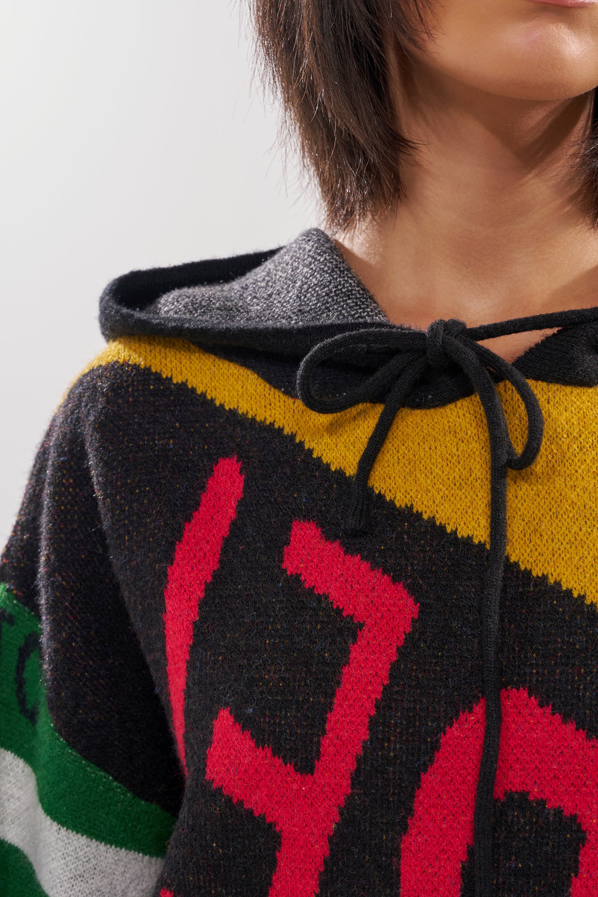 A close-up shows a person wearing the ALPHA FEMALE HOODED SWEATSHIRT DRESS IN YELLOW MULTI, featuring a bold yellow, green, red, and black patchwork design with a hood and black drawstrings. Part of their short dark hair is visible.