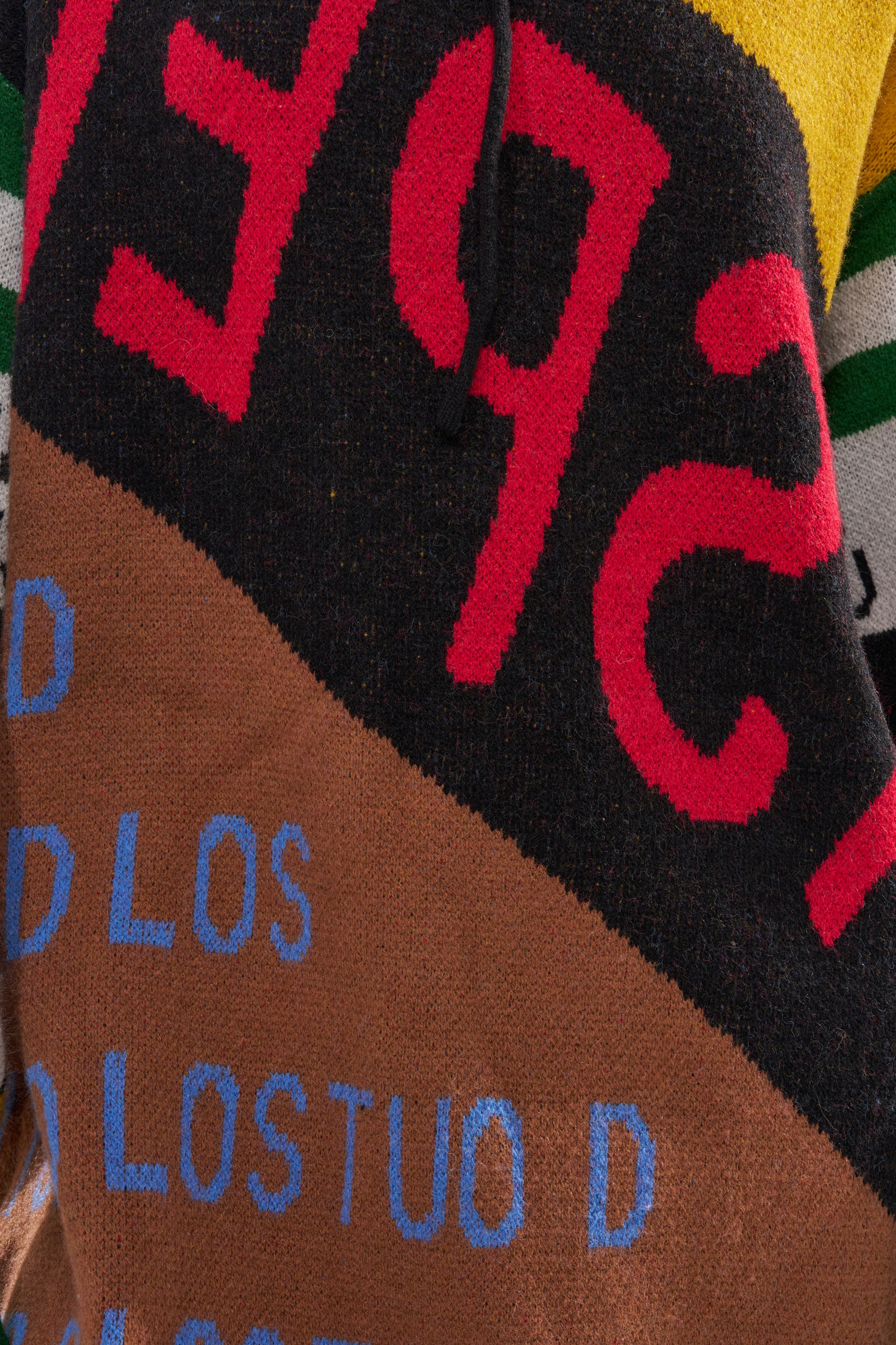 A close-up of the ALPHA FEMALE HOODED SWEATSHIRT DRESS IN YELLOW MULTI shows colorful knitted fabric with abstract shapes and bold "LOSTUO D" text in blue, reflecting a vibrant patchwork streetwear-inspired design.