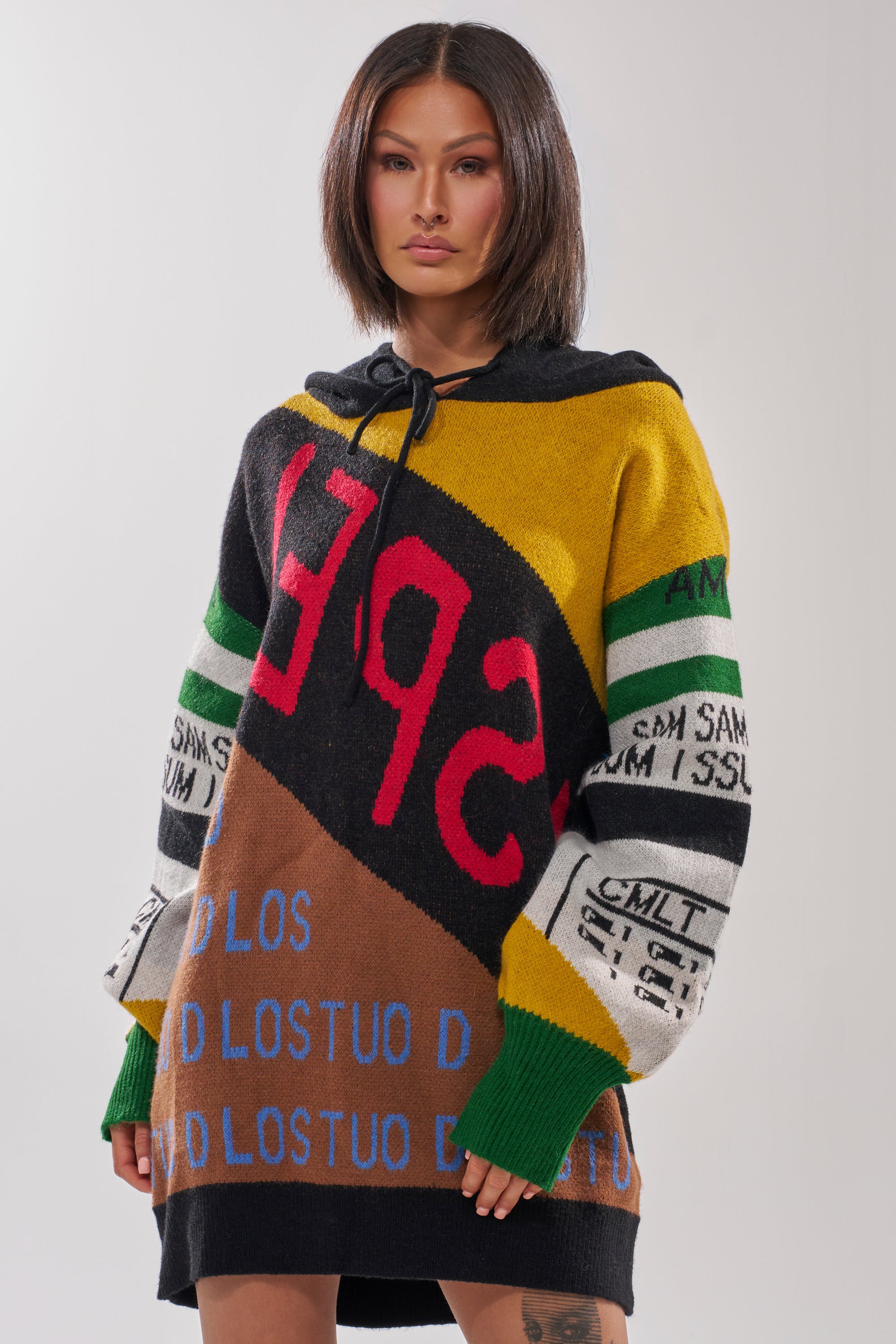 A woman with straight dark hair wears the ALPHA FEMALE HOODED SWEATSHIRT DRESS IN YELLOW MULTI, a bold oversized streetwear piece with colorful graphic text accents, standing against a plain light background.