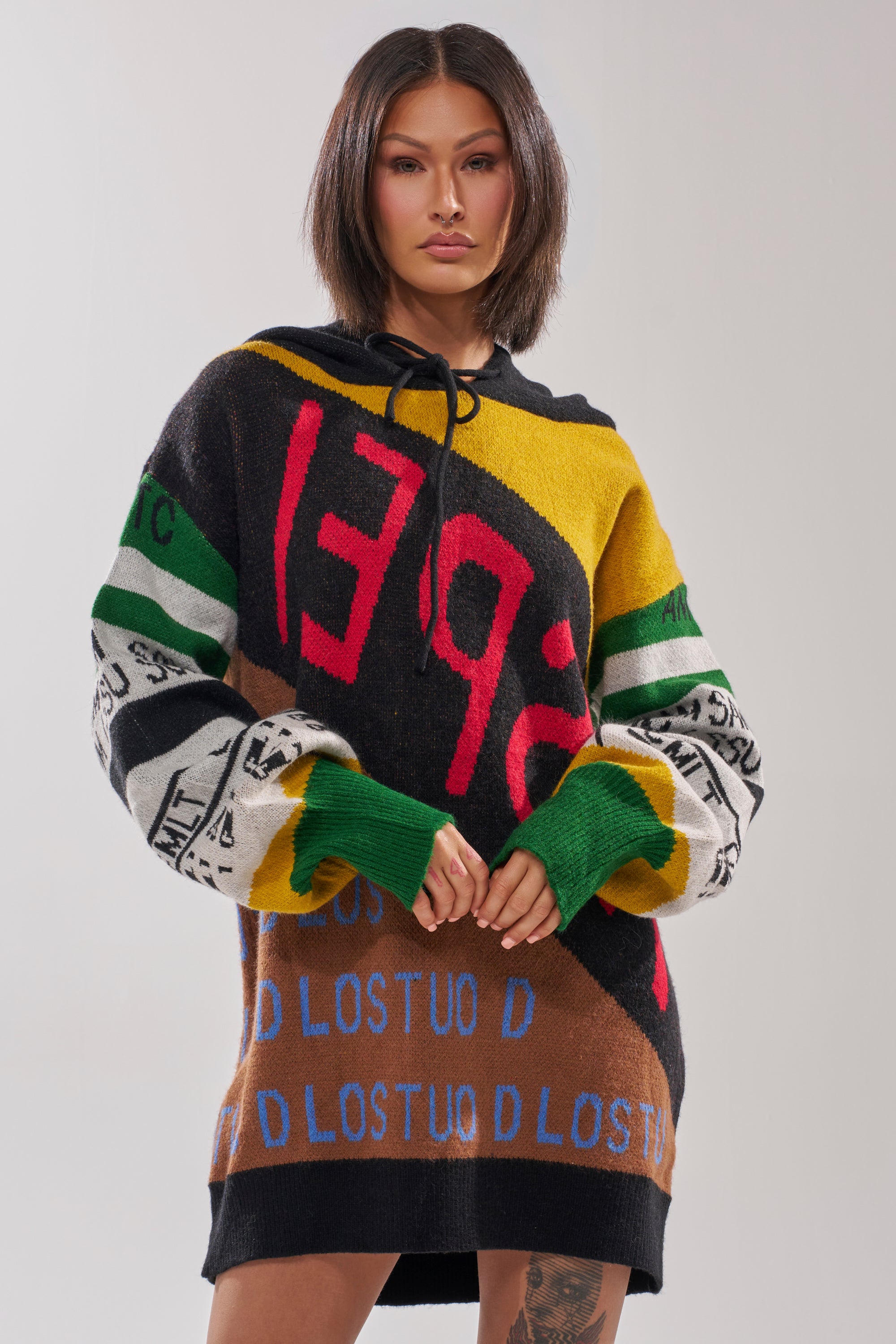 A woman with shoulder-length dark hair wears the ALPHA FEMALE HOODED SWEATSHIRT DRESS IN YELLOW MULTI, featuring colorful abstract patterns and bold, mixed lettering. She stands facing forward against a plain light background.
