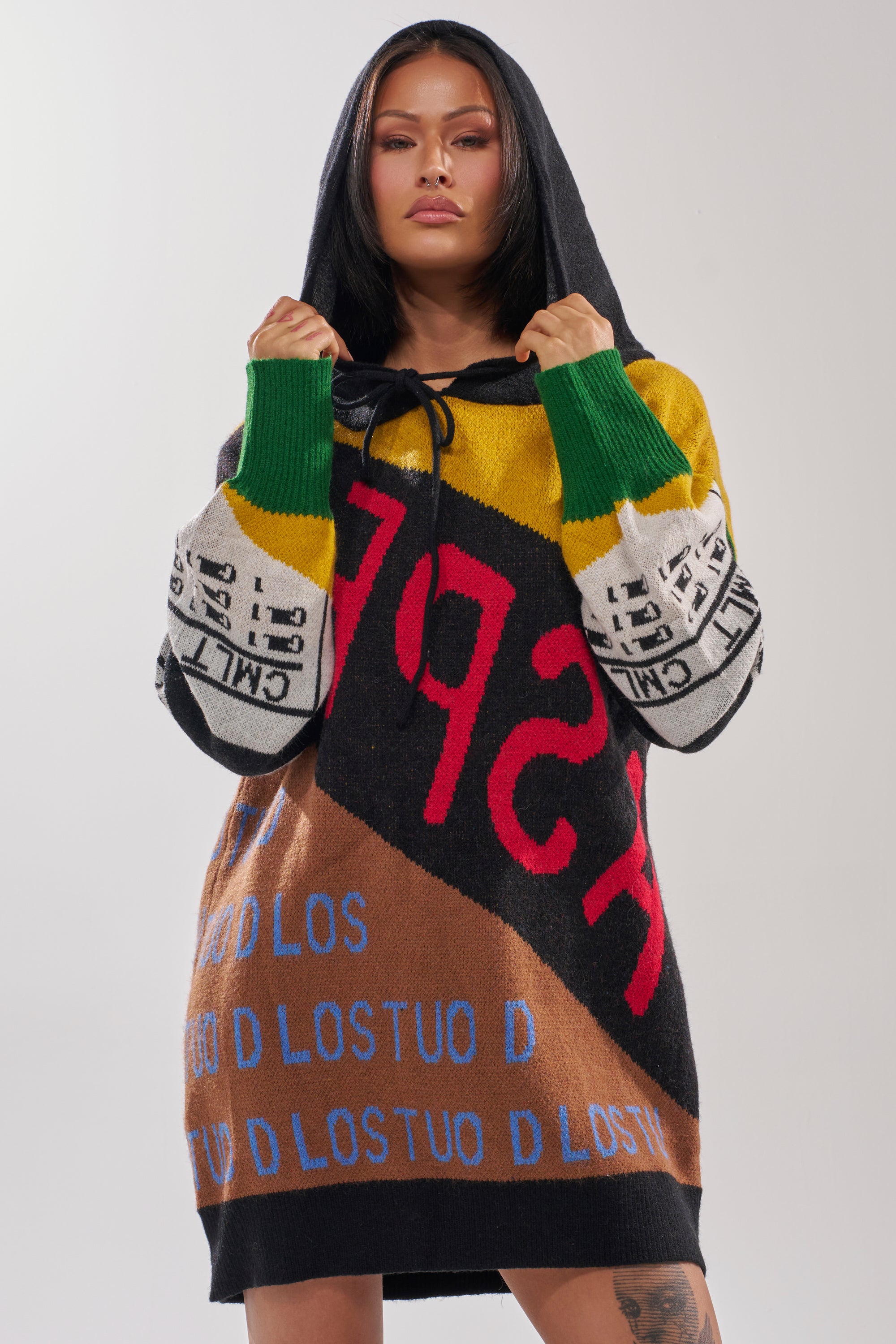 A woman with straight dark hair wears the ALPHA FEMALE HOODED SWEATSHIRT DRESS IN YELLOW MULTI, featuring colorful patchwork, large letters, and repeating text. She faces forward and holds the hood's drawstrings.