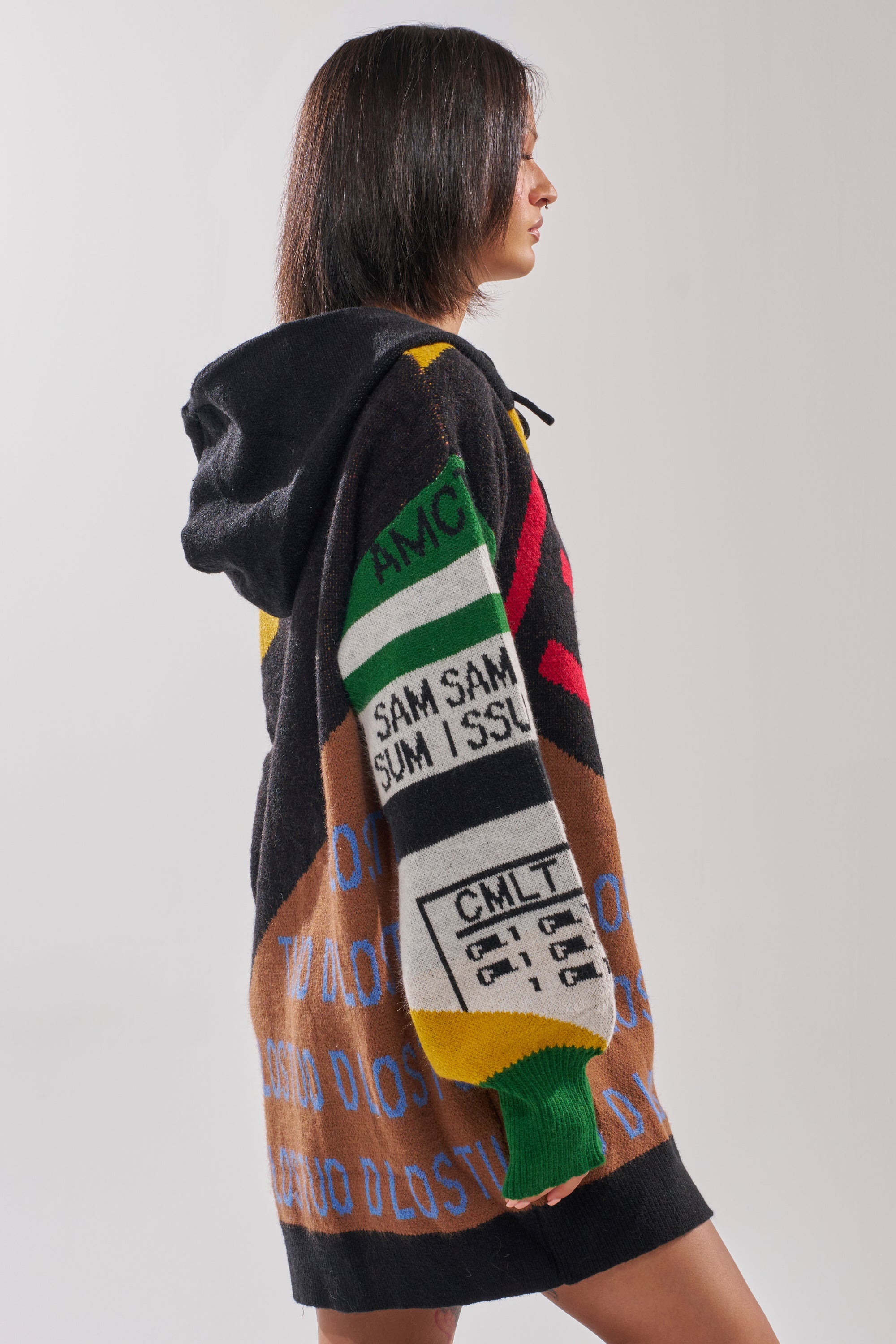 A person with shoulder-length dark hair stands sideways, wearing the ALPHA FEMALE HOODED SWEATSHIRT DRESS IN YELLOW MULTI, which features colorful geometric patterns and bold text blocks on the back.