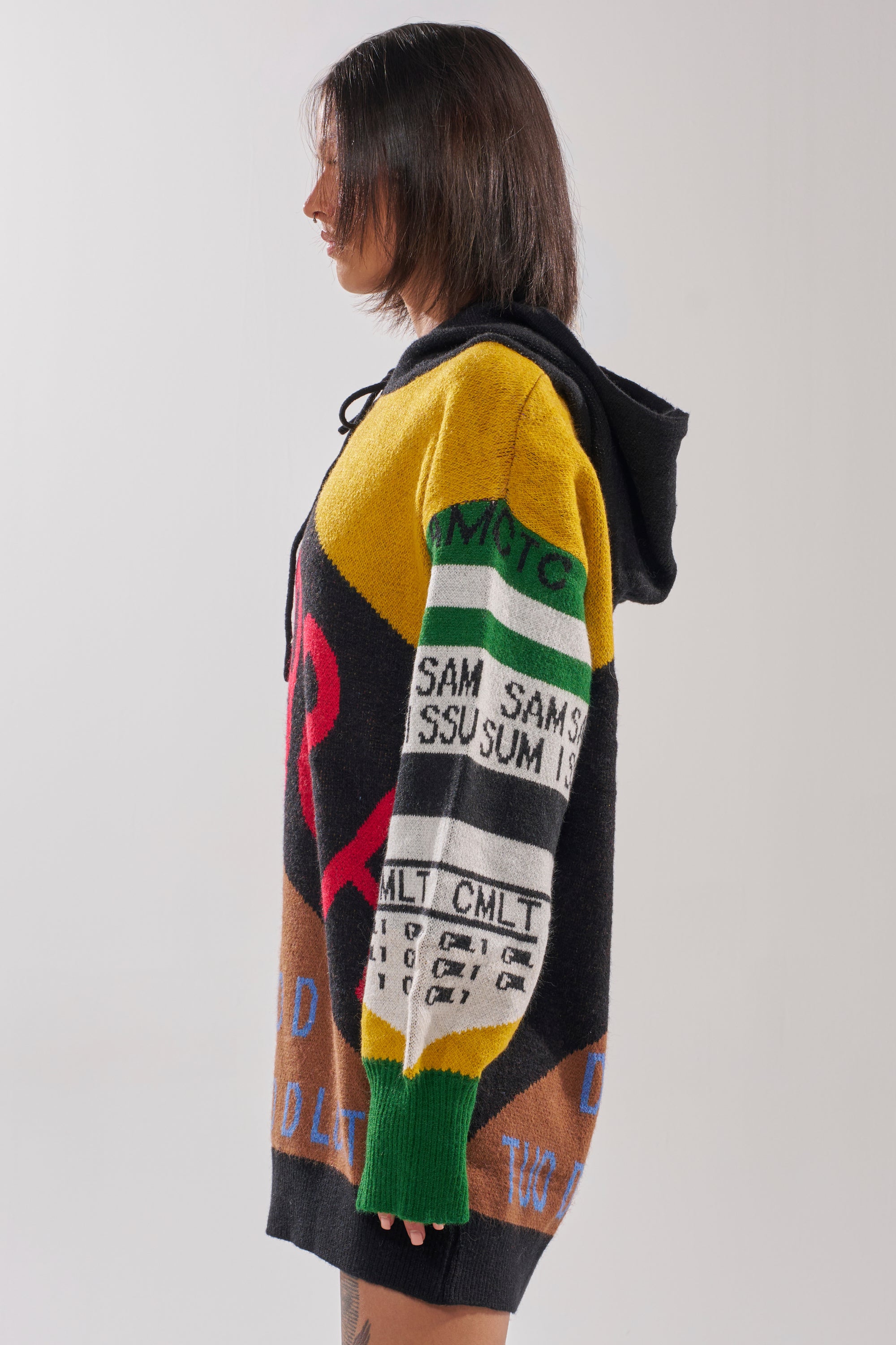 A person with straight, shoulder-length hair stands in profile, wearing the ALPHA FEMALE HOODED SWEATSHIRT DRESS IN YELLOW MULTI, which features a vibrant patchwork design with graphic text and bold patterns in yellow, green, black, and red.