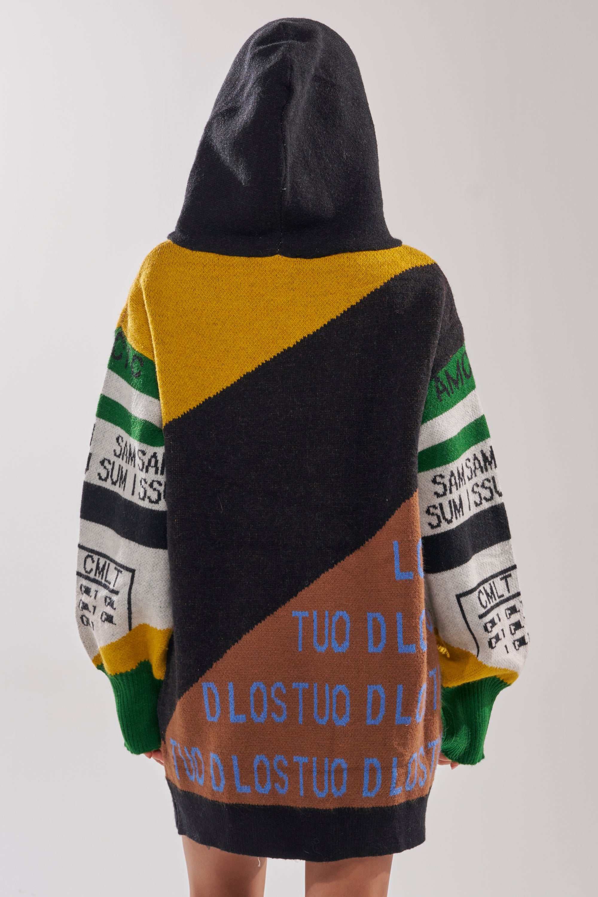 Wearing the ALPHA FEMALE HOODED SWEATSHIRT DRESS IN YELLOW MULTI, a person faces away, showing bold geometric patchwork in yellow, black, brown, and green with “SAMSUMISS” and “D LOSTU” on the back and sleeves.