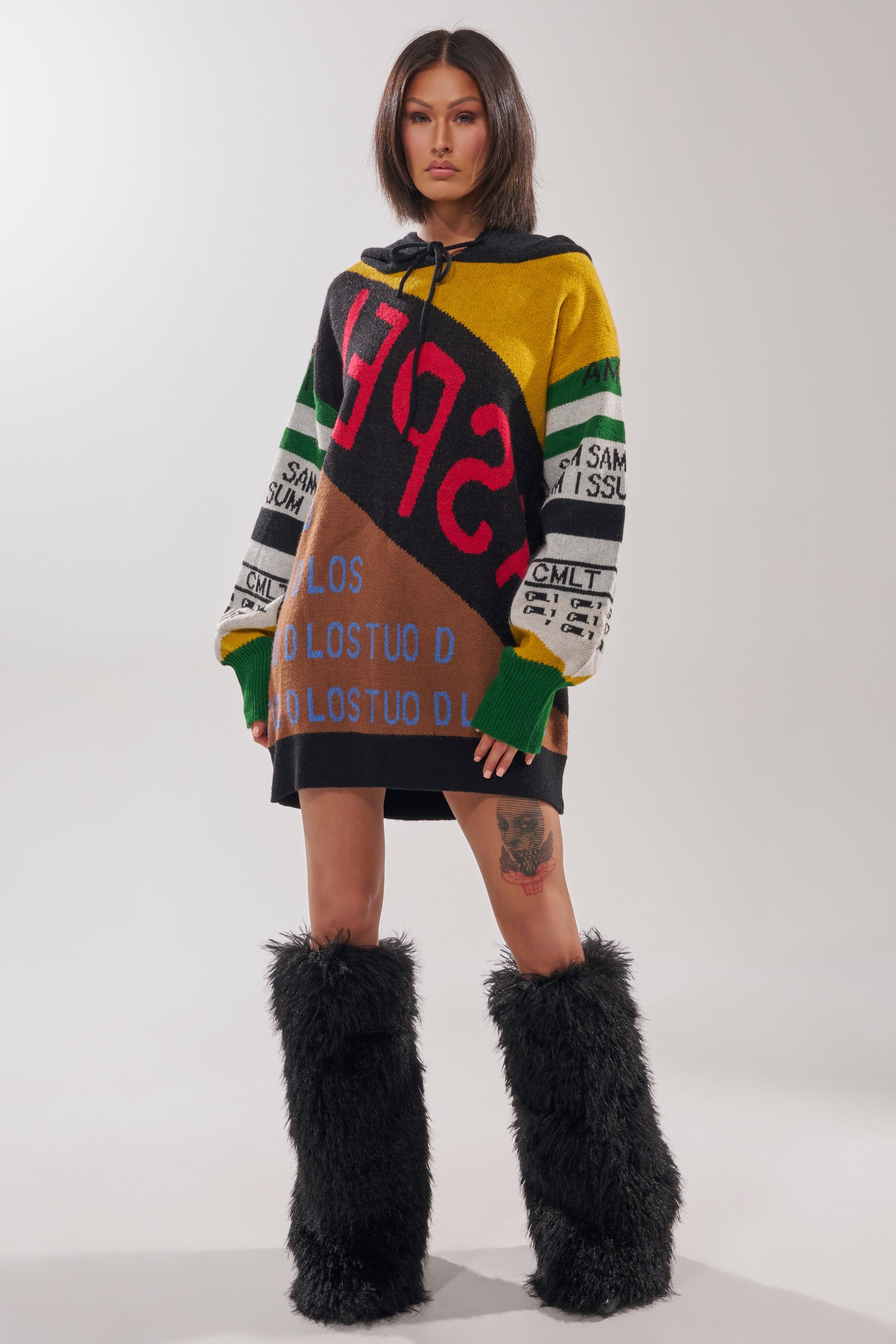 A woman with straight dark hair models the ALPHA FEMALE HOODED SWEATSHIRT DRESS IN YELLOW MULTI, featuring a bold patchwork design and graphic text, paired with black furry knee-high boots against a plain white background.