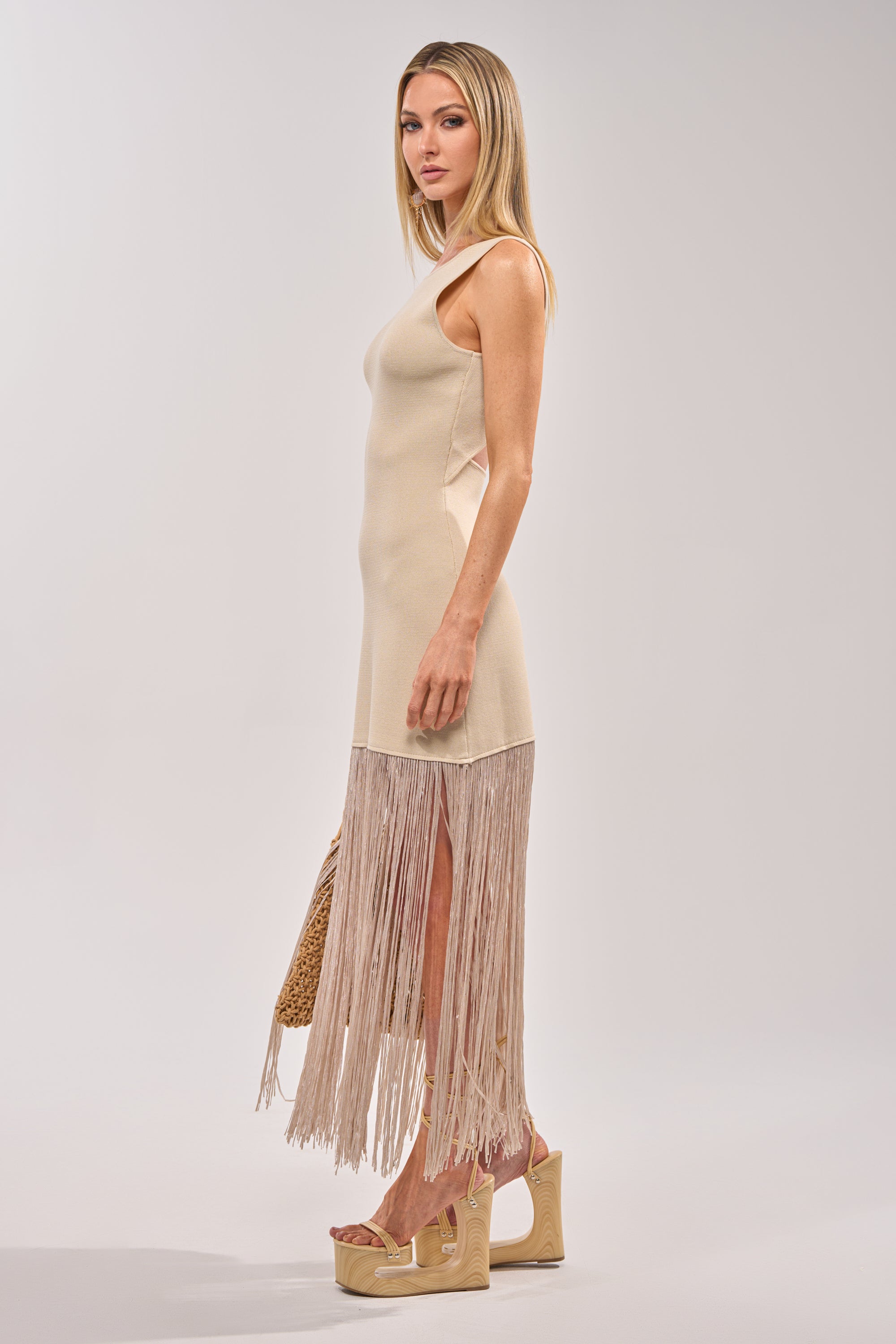 A woman stands in profile wearing the FLY AWAY KNIT MIDI DRESS WITH FRINGE, a sleeveless, fitted beige cocktail dress with a fringe hemline. She holds a woven bag and wears high platform sandals against a plain light background. 3