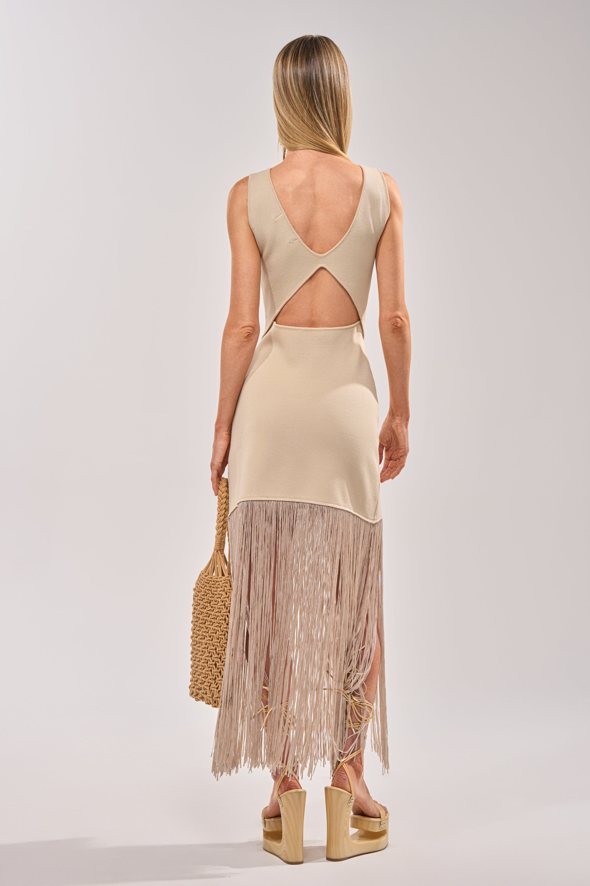 A woman with straight blonde hair is seen from behind wearing the FLY AWAY KNIT MIDI DRESS WITH FRINGE, a sleeveless beige cocktail dress featuring a back cutout and long fringe hem, styled with a woven straw handbag and beige platform sandals. 4