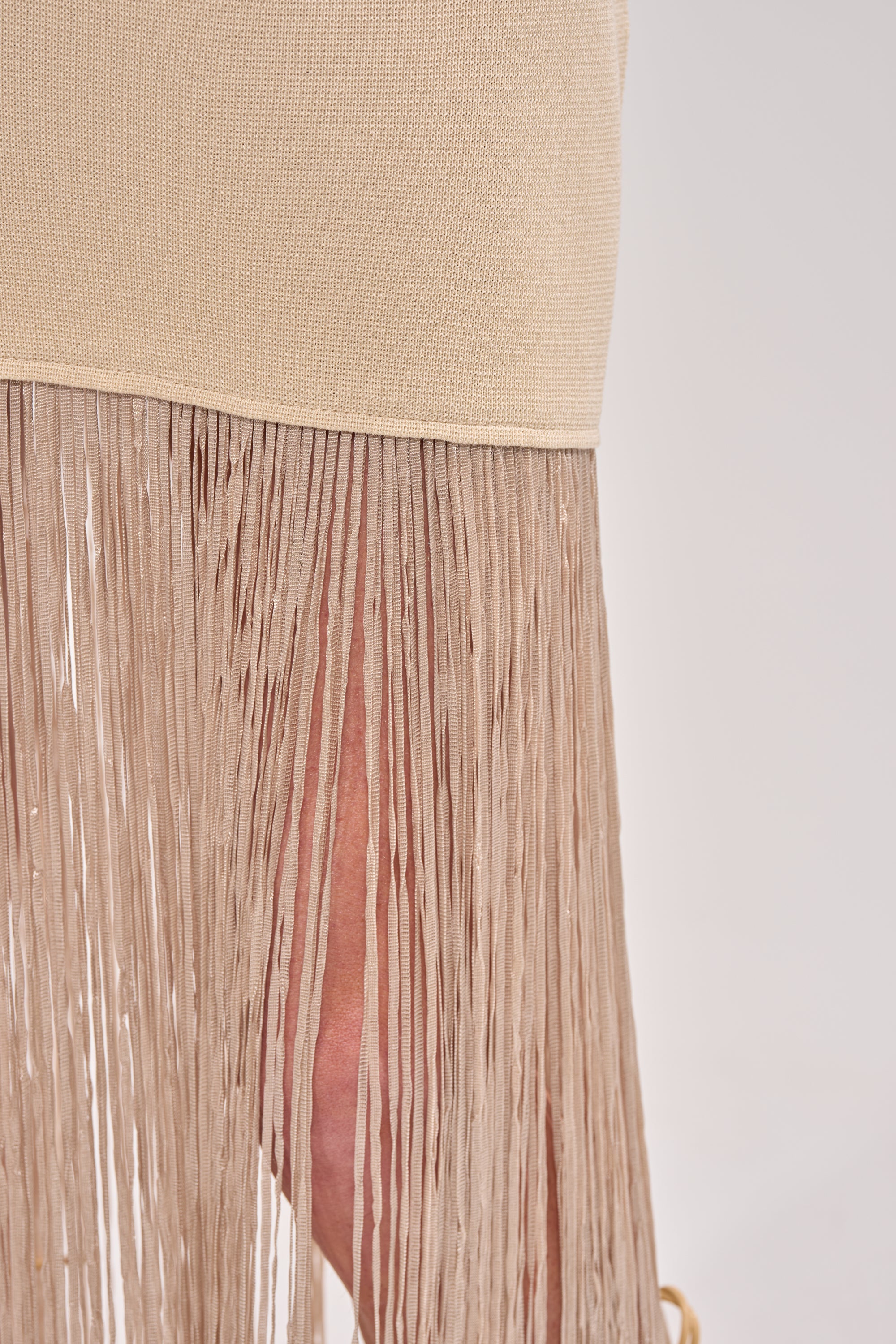 Close-up of the FLY AWAY KNIT MIDI DRESS WITH FRINGE in beige, featuring a long, thin fringe hemline that reveals skin beneath. Set against a plain, light-colored background. 5