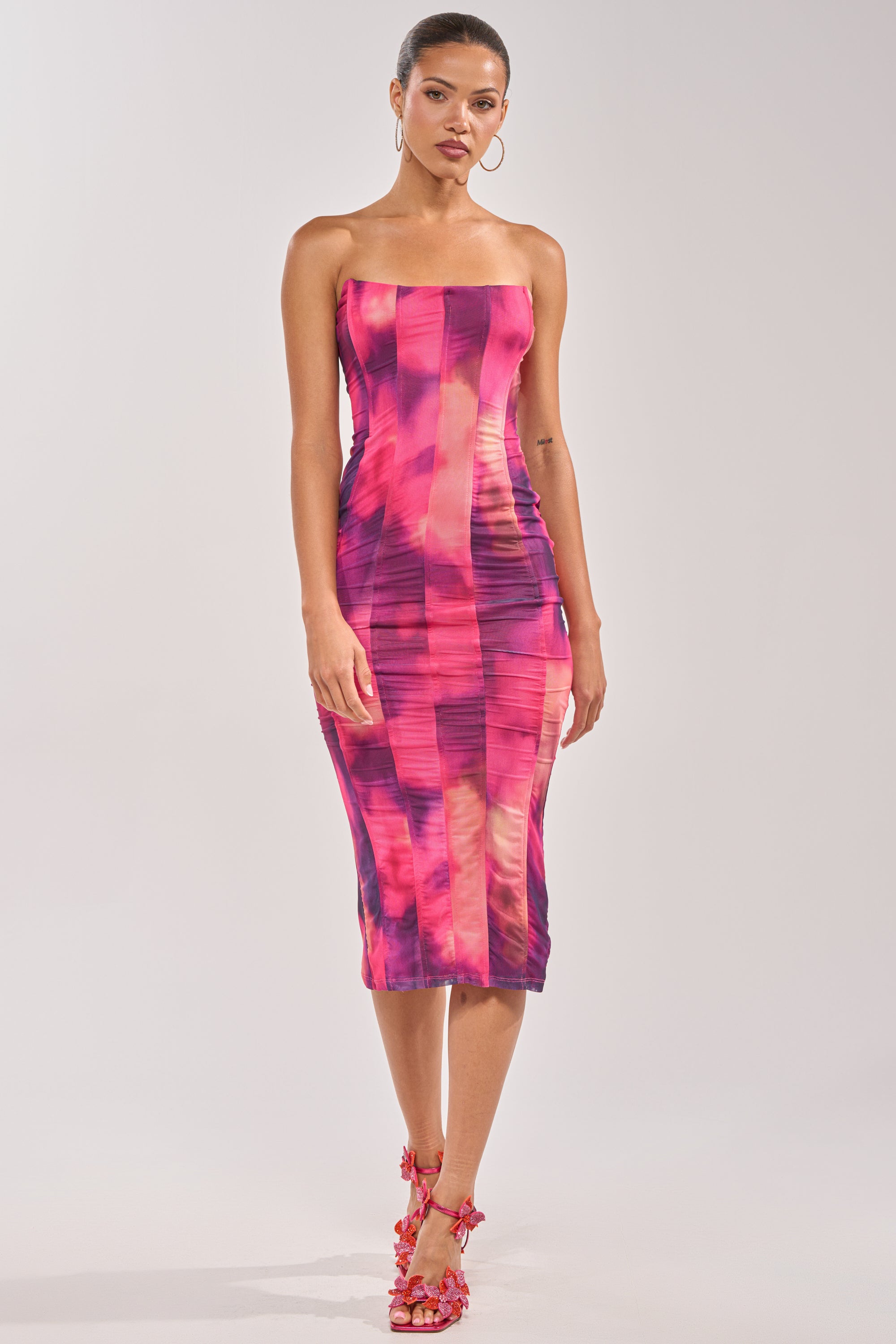 A woman stands against a plain background wearing the ROSE GARDEN MAXI DRESS IN PINK MULTI, a strapless, figure-hugging dress with a pink and purple geometric pattern. She pairs it with matching pink heels, hoop earrings, and has her hair pulled back. 0