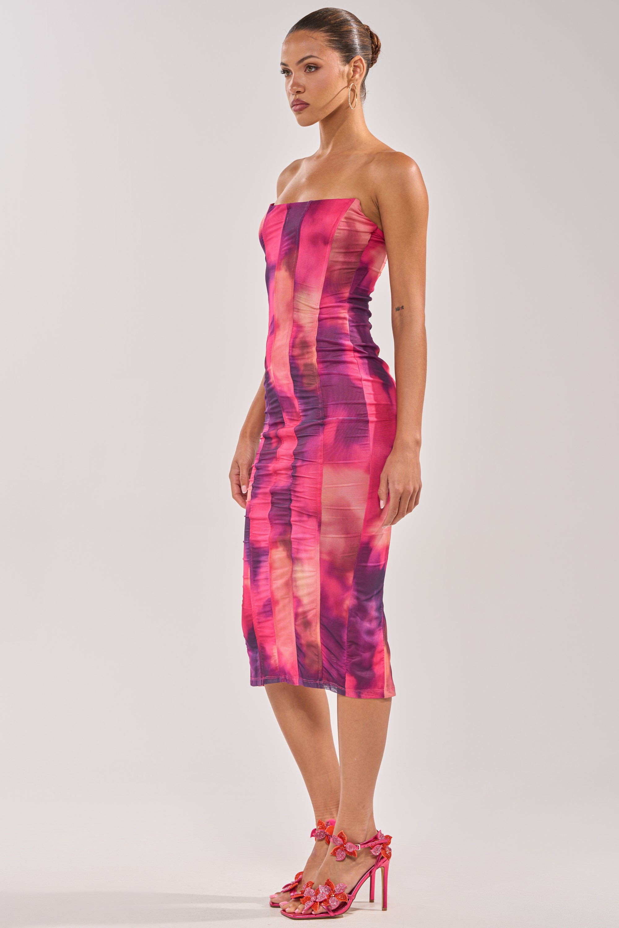 A woman stands sideways in the ROSE GARDEN MAXI DRESS IN PINK MULTI, a strapless, body-hugging evening dress with a pink and purple abstract pattern. She pairs it with matching floral high-heeled sandals and wears her hair in a neat bun. 2