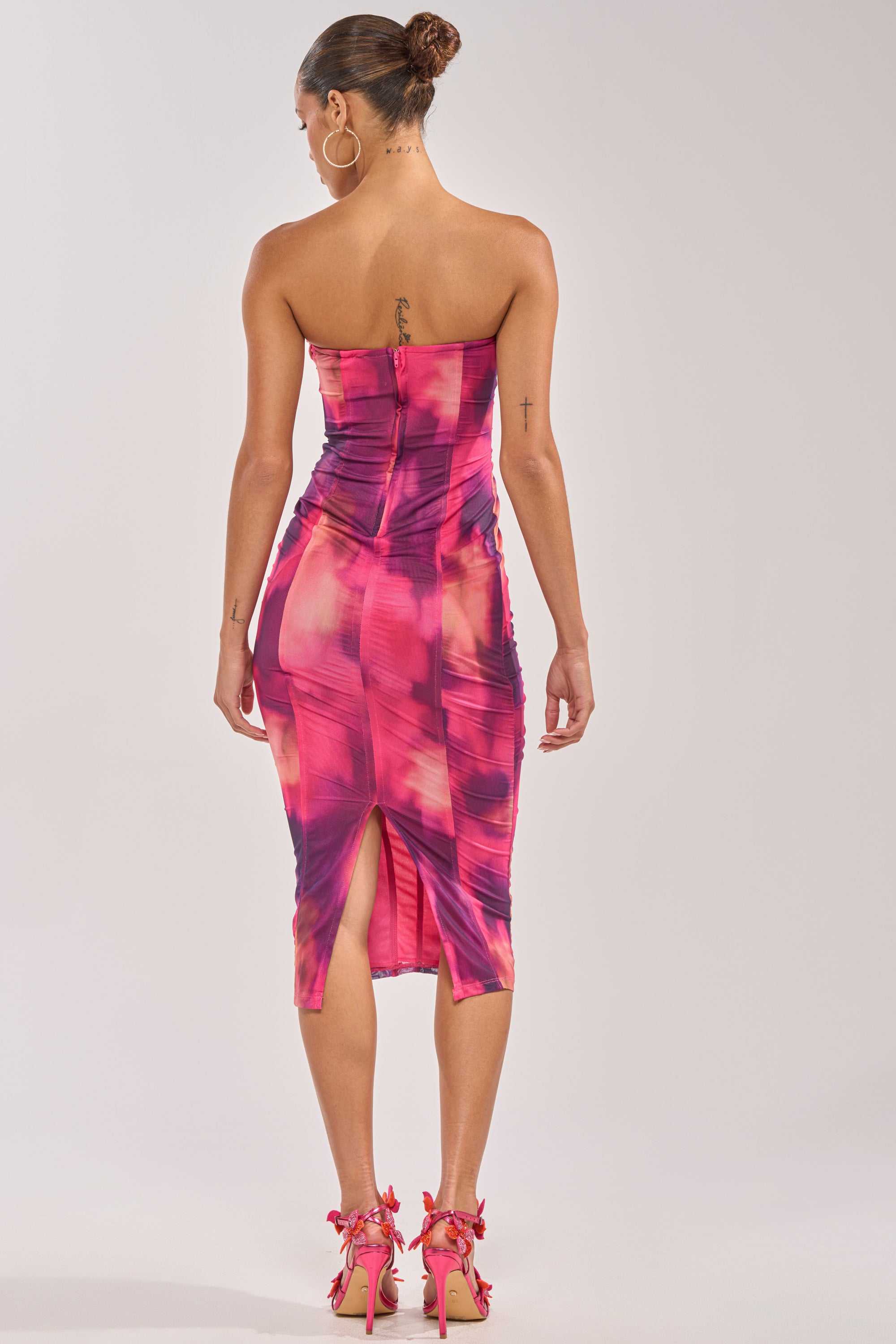 A woman with a tattoo on her upper back wears the ROSE GARDEN MAXI DRESS IN PINK MULTI, a strapless, body-hugging style with pink and purple patterns, back slit, and matching heels. She stands facing away from the camera. 3