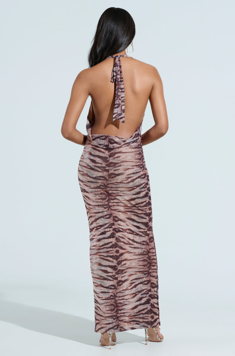 A woman with long dark hair stuns in a FIERCE ZEBRA MAXI DRESS, showcasing a backless halter-neck style and striking tiger stripe pattern. The ankle-length dress pairs perfectly with her strappy high-heeled sandals as she stands against a light backdrop, showing off the dress's elegance.