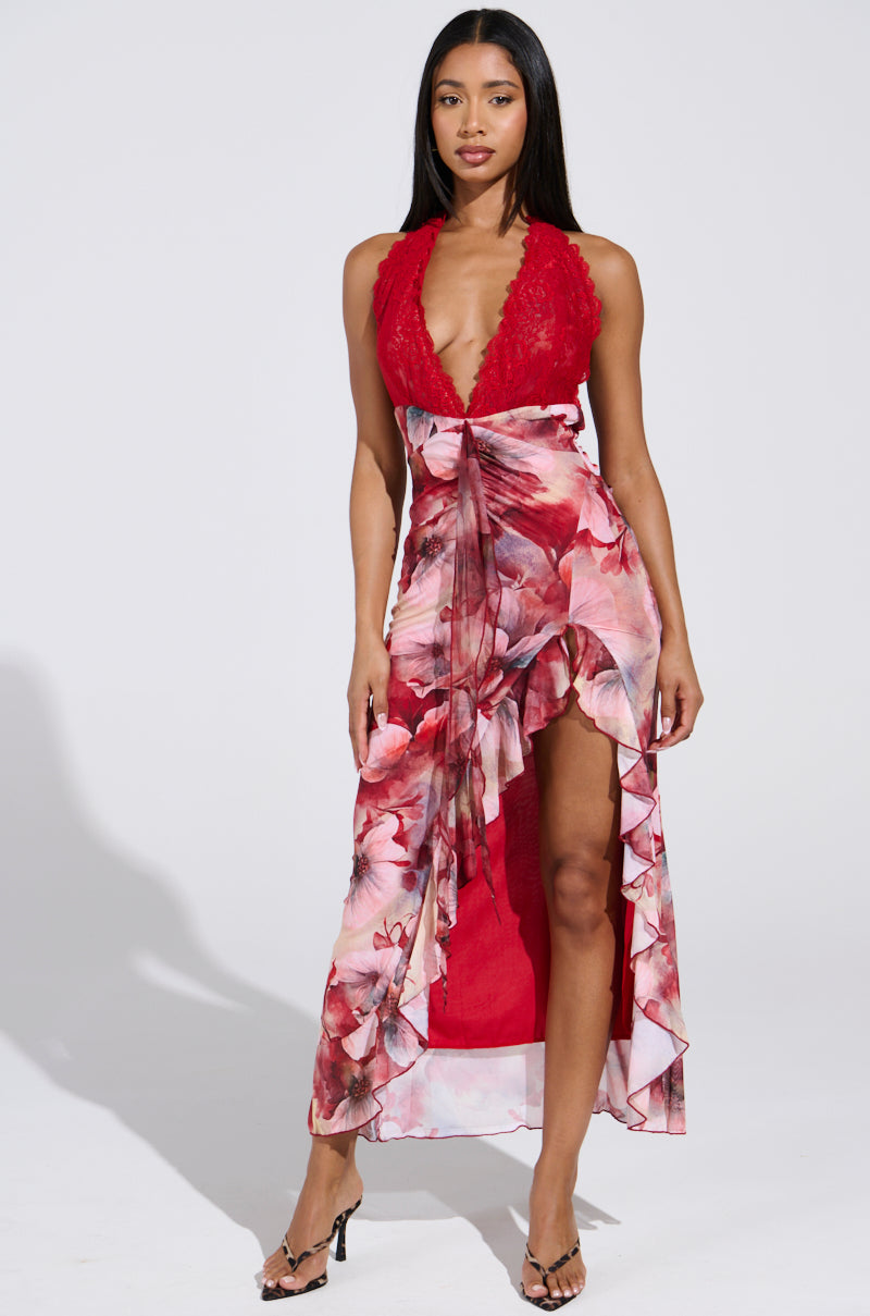 Against a plain background, someone poses in the ALIA FLORAL MIDI DRESS, showcasing its vibrant floral pattern with a red lace top and a flowing skirt. The dress features a high slit and a ruffle-trimmed design. Their long hair complements the elegant look.