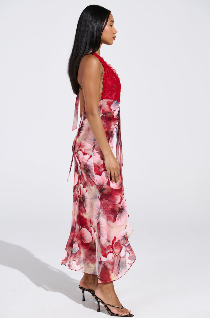 In profile against a white background, a woman with long, straight hair and high-heeled sandals showcases the ALIA FLORAL MIDI DRESS. This red midi dress features pink and red floral patterns, lace accents, and a ruffle-trimmed flowing skirt.