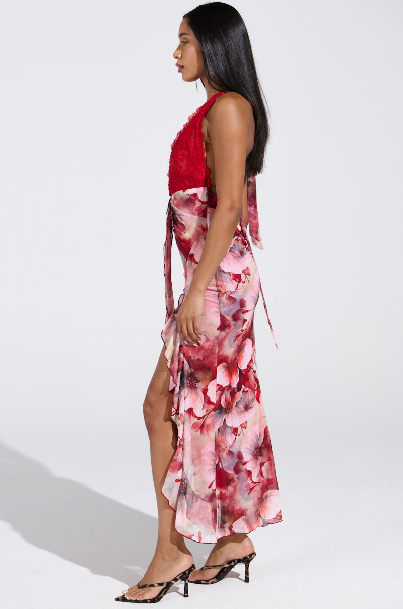 In a plain, well-lit setting, a woman with long hair is in profile wearing the ALIA FLORAL MIDI DRESS and open-toed heels.