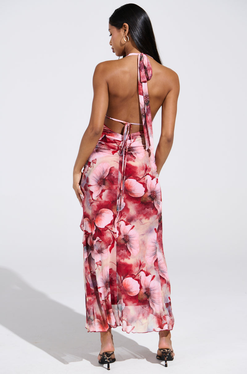 Wearing the ALIA FLORAL MIDI DRESS, a woman with long hair poses elegantly in high heels. This dress features a halter neckline, high slit, open back with ties, and is adorned in shades of pink, red, and white floral patterns. She stands facing away from the camera.