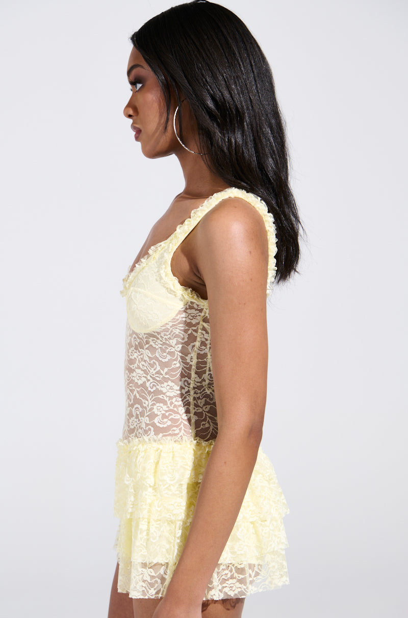In profile against a white background, the woman wears a DREAMING ABOUT YOU LACE MINI DRESS with yellow floral lace and ruffled layers. Her long dark hair cascades down, complementing large hoop earrings and the dress's curve-hugging silhouette.