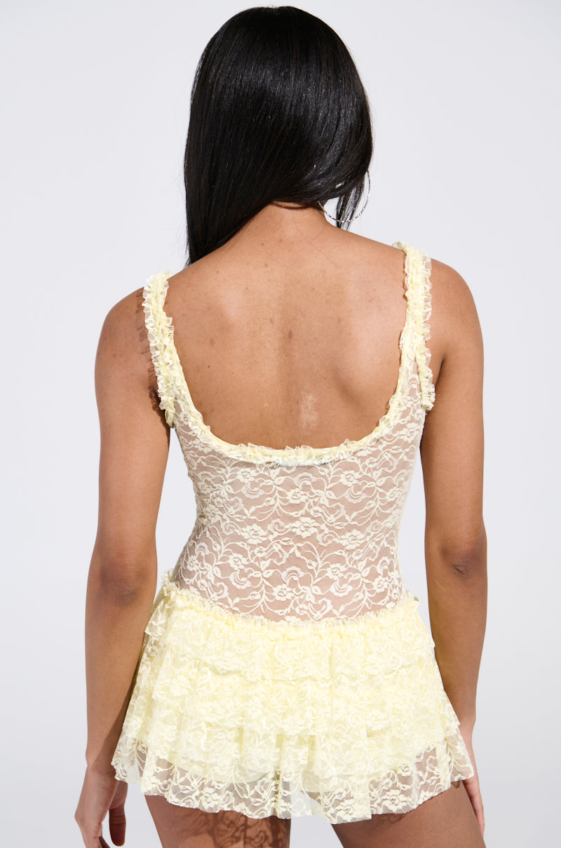 Rear view of a person with long black hair wearing a sheer yellow DREAMING ABOUT YOU Lace Mini Dress, highlighting its ruffled layers and curve-enhancing fit against a plain white background.