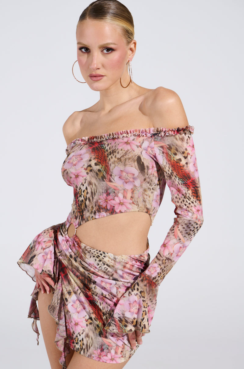 Wearing the MIAMI NIGHTS MINI DRESS, featuring floral and animal prints with a one-shoulder top and matching skirt, a person exudes confidence. Paired with hoop earrings and slicked-back hair, they radiate chic elegance against a neutral backdrop.