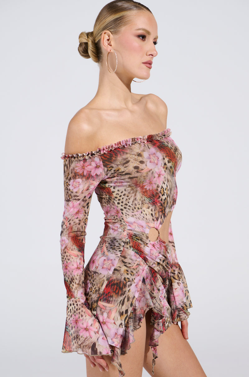 A person wears the MIAMI NIGHTS MINI DRESS, an off-the-shoulder piece with pink floral and animal prints, featuring a ruffled neckline and hem plus a cutout waist. Their hair is tied back and accessorized with large hoop earrings.