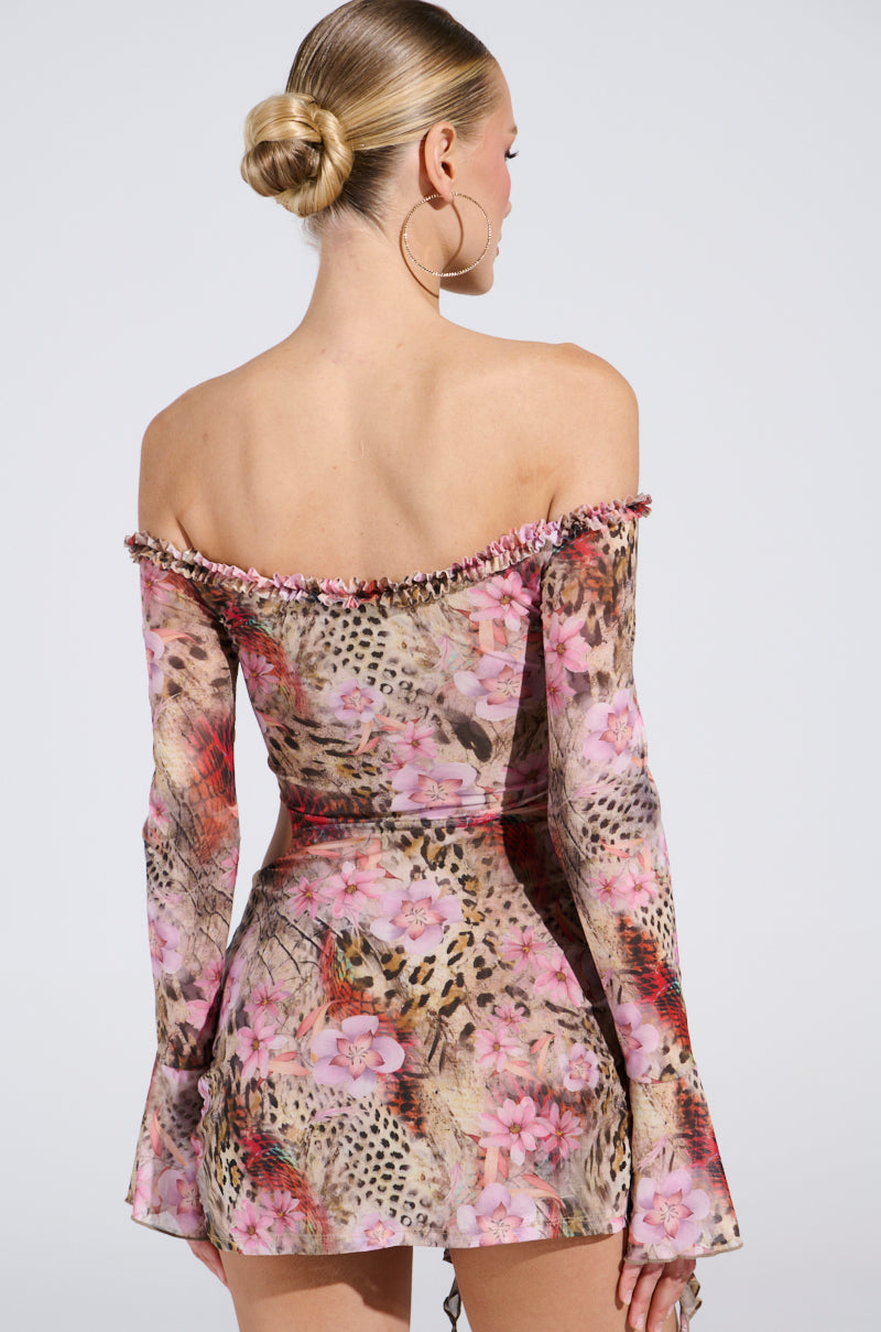 Wearing the Miami Nights Mini Dress, a person stands with their back to the camera. The dress boasts an off-the-shoulder design, floral and leopard print, and long sleeves. They have styled hair in a bun and large hoop earrings.