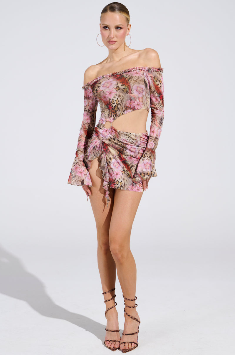 Dazzling in a floral and animal print MIAMI NIGHTS MINI DRESS with an off-the-shoulder neckline, cutouts, and a ruffled skirt, the person completes the look with large hoop earrings and strappy high heels against a plain white background.